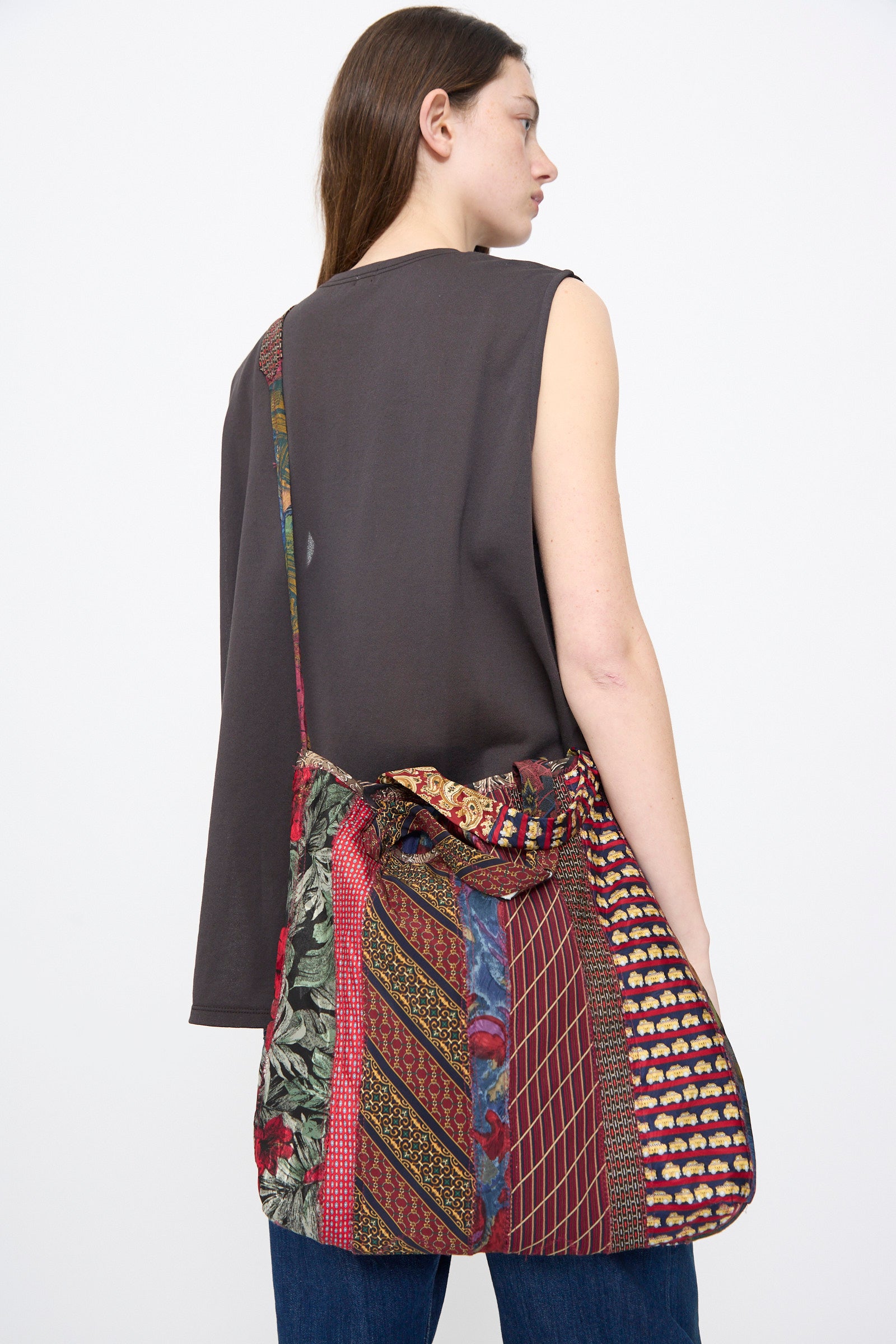 A woman in a sleeveless dark top and jeans stands with her back turned, carrying the Intensity Ties Bag in Red Multi—a large, unique patchwork silk tie bag—over her shoulder.