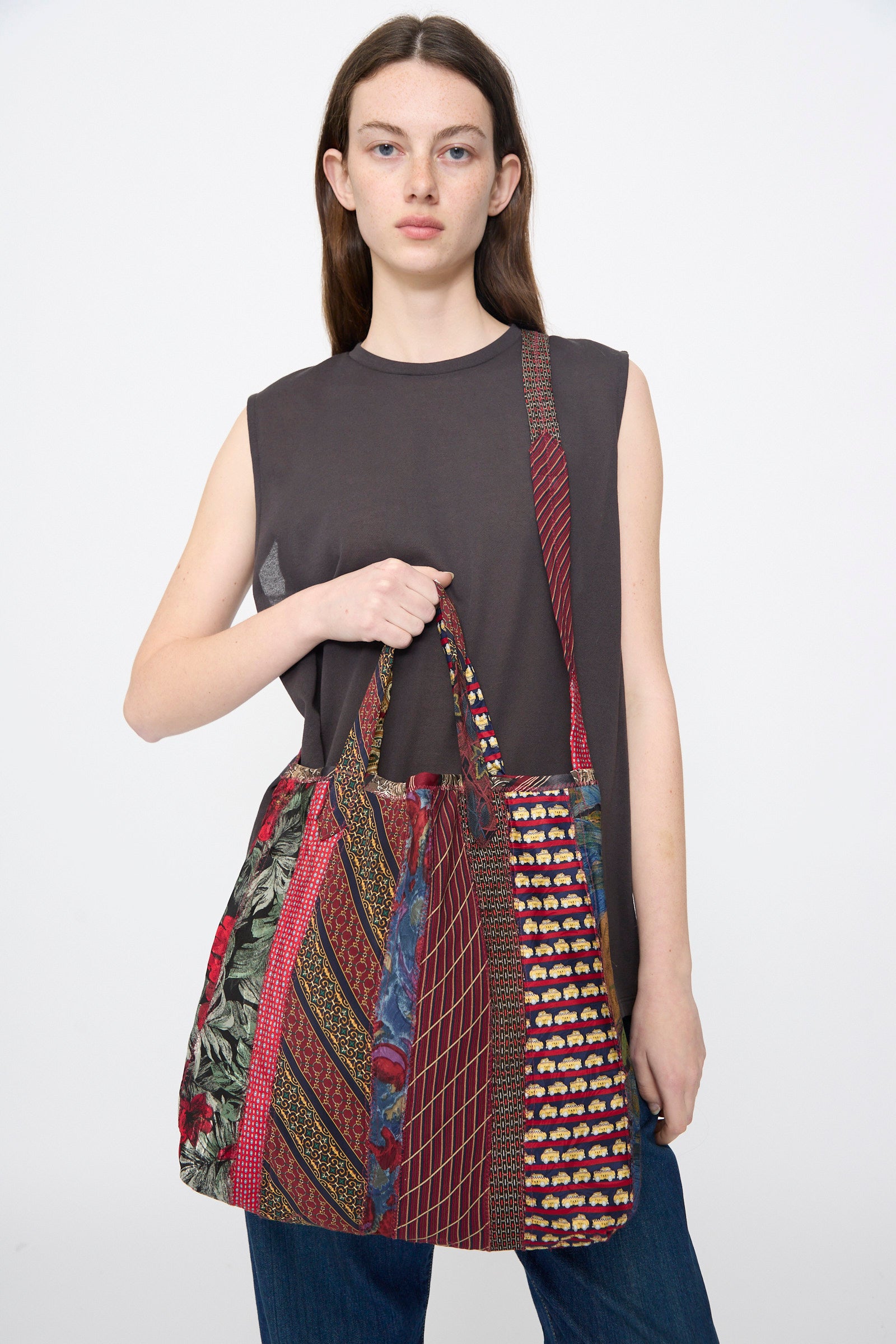A woman with straight brown hair wears a sleeveless dark gray top and jeans, carrying the Intensity Ties Bag in Red Multi—a large, colorful patchwork silk tie tote.