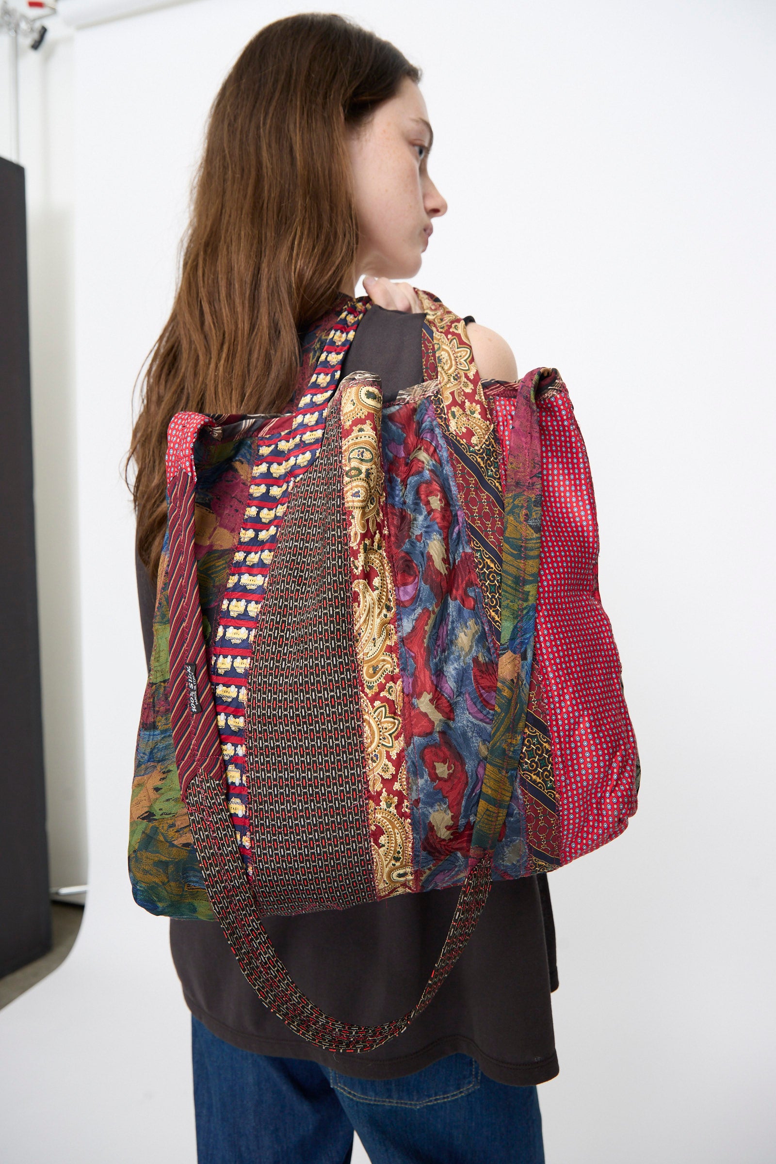 A woman with long brown hair in a dark top and blue jeans carries the Intensity Ties Bag in Red Multi, a large, colorful patchwork tote, over her shoulder.