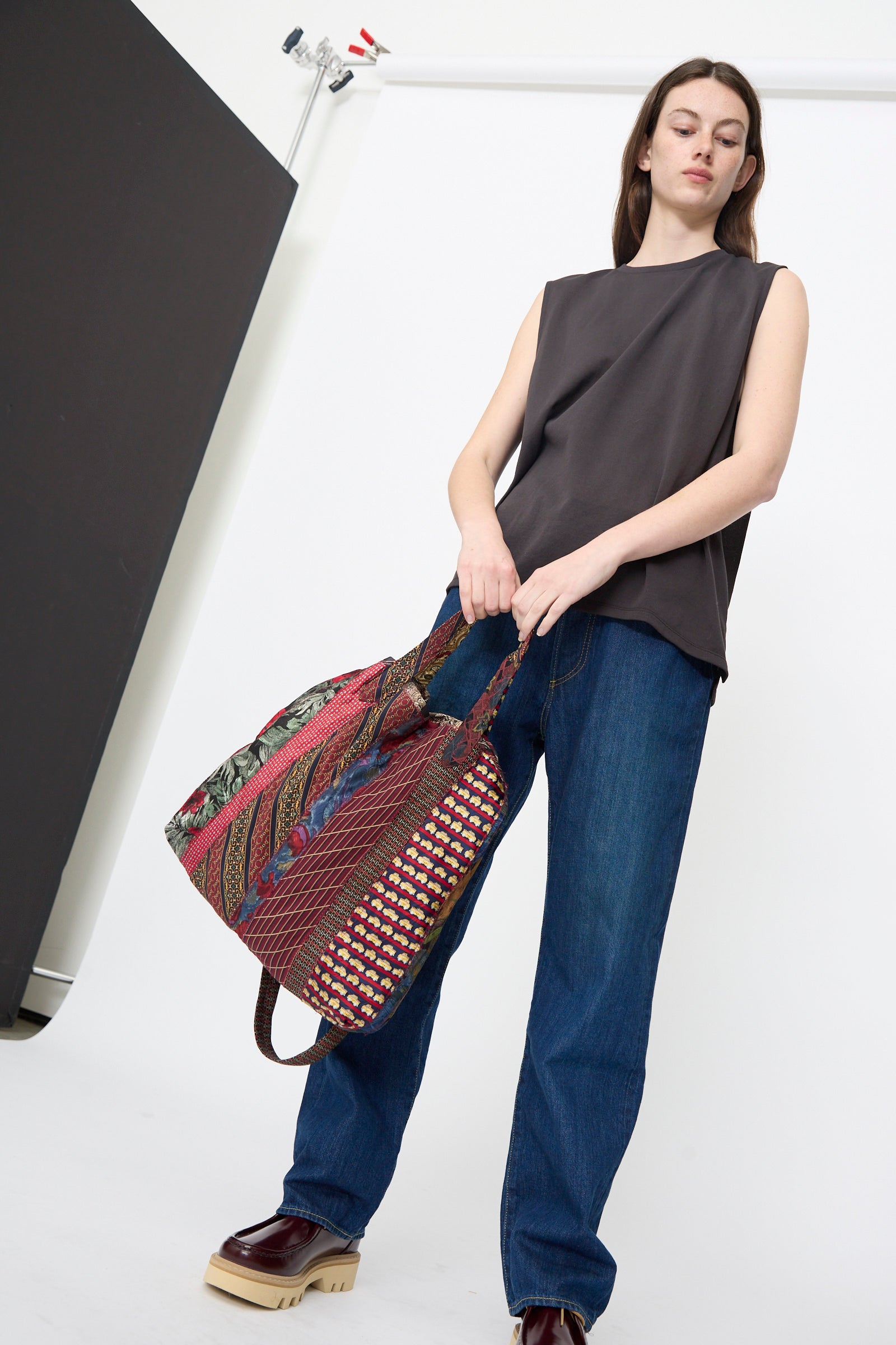 A person wearing a sleeveless dark top, blue jeans, and platform shoes stands in a minimalist studio holding the Intensity Ties Bag in Red Multi.