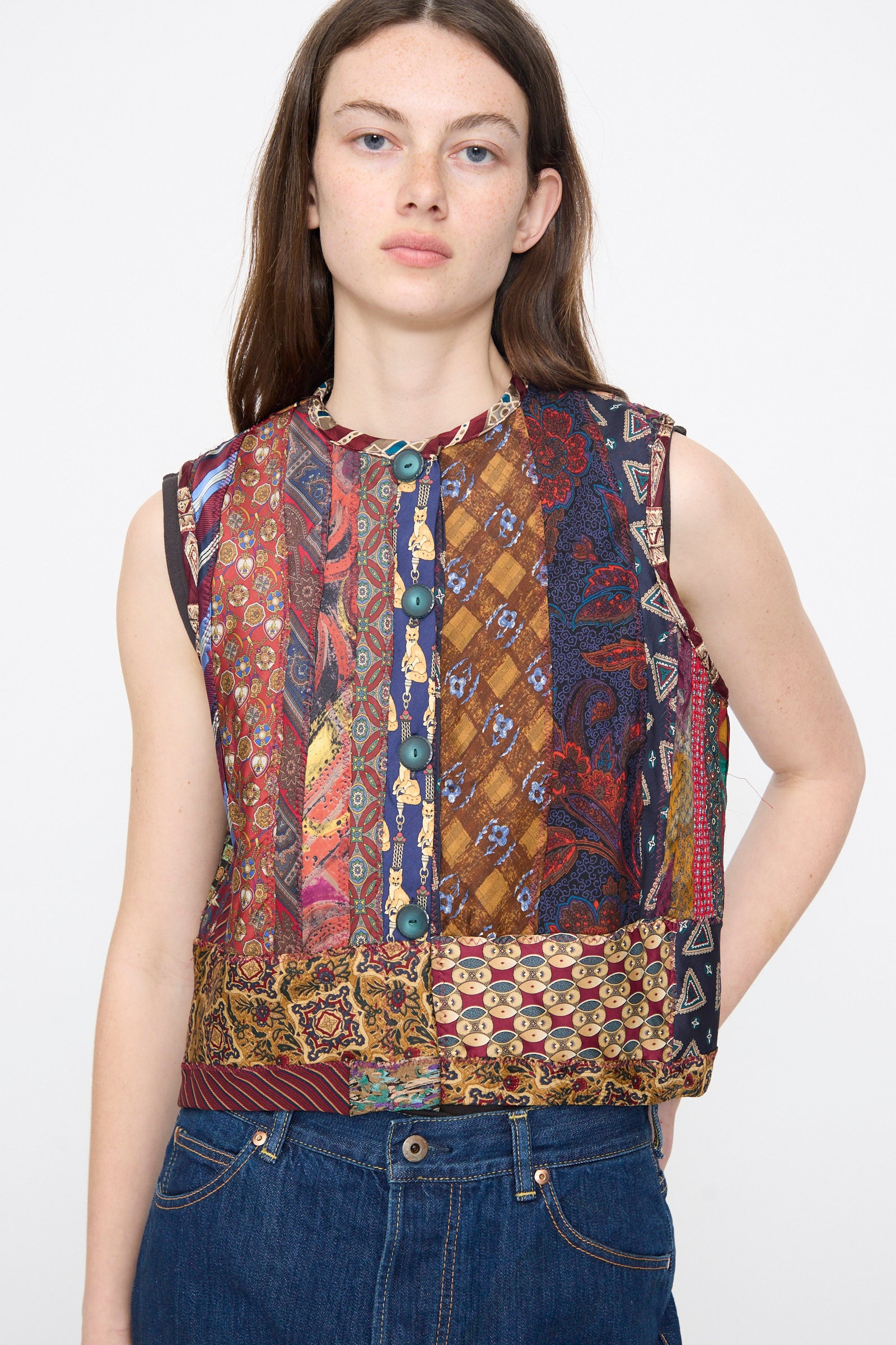 A person wearing the Intensity Ties Vest in Multi—a sleeveless, multicolored patchwork silk tie vest with blue buttons—and dark blue jeans stands against a plain light background.