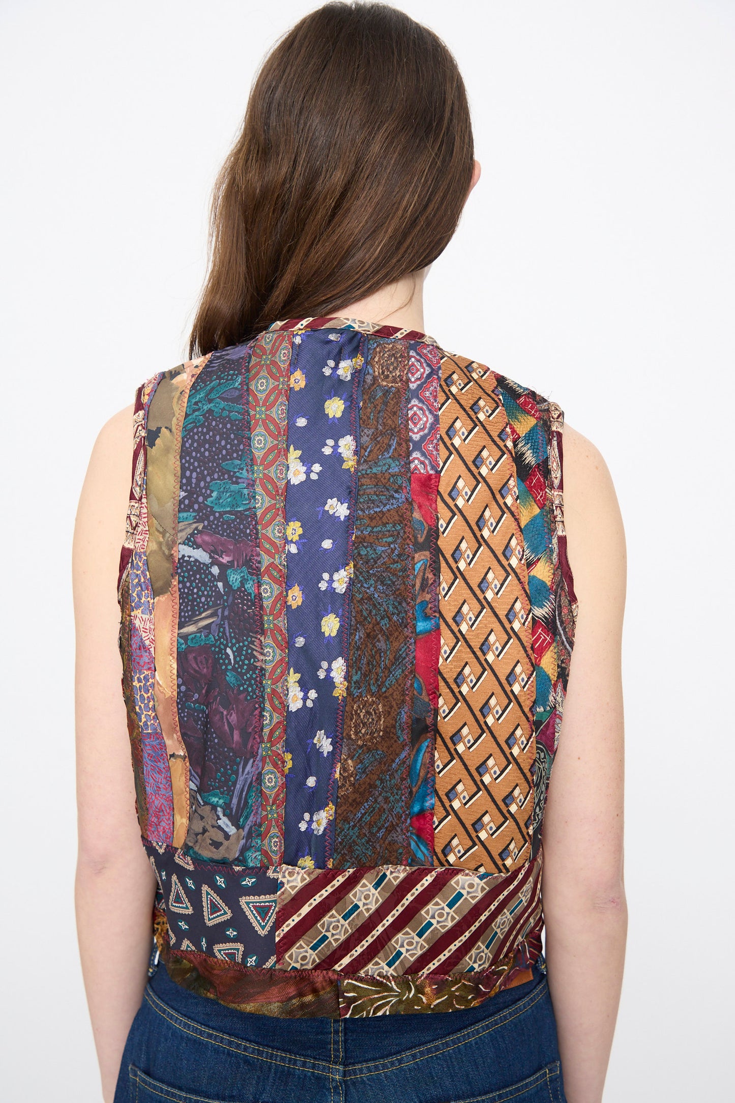 A person with long brown hair, shown from the back, wears the Intensity Ties Vest in Multi—a sleeveless, one-of-a-kind vest featuring a patchwork of colorful patterns.