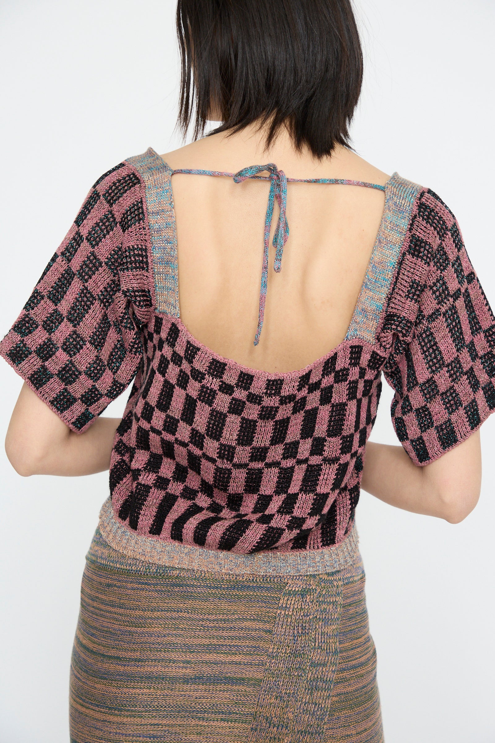 A model seen from the back wearing the Wing Corset in Pink by Intensity, with  a checkered pink and black design, lace-up closure, tied around the neck.