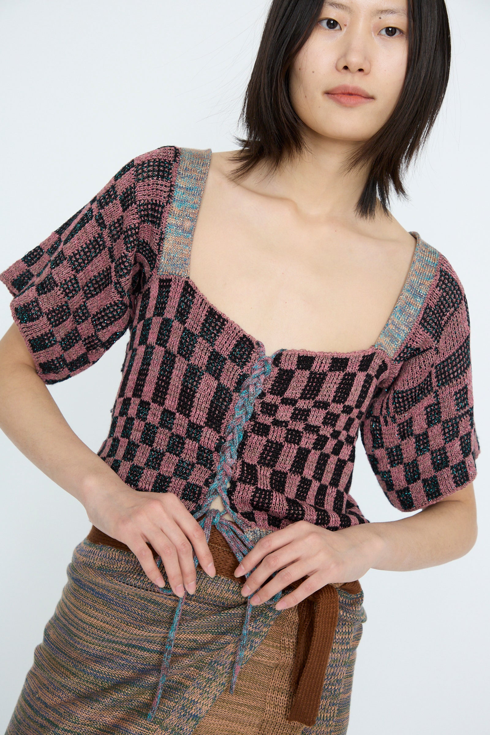 A model is wearing the Wing Corset in pink by Intensity, featuring a checkered pattern and lace-up closure, along with a textured skirt woven with metallic yarn against a white backdrop.