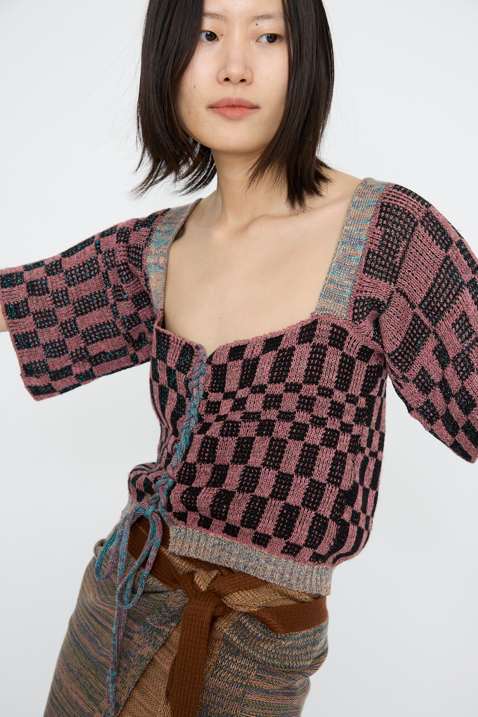 A person wears Intensity's Wing Corset in Pink, featuring a pink and black checkered design with a square neckline and lace-up closure, paired with a brown skirt against a white backdrop. 