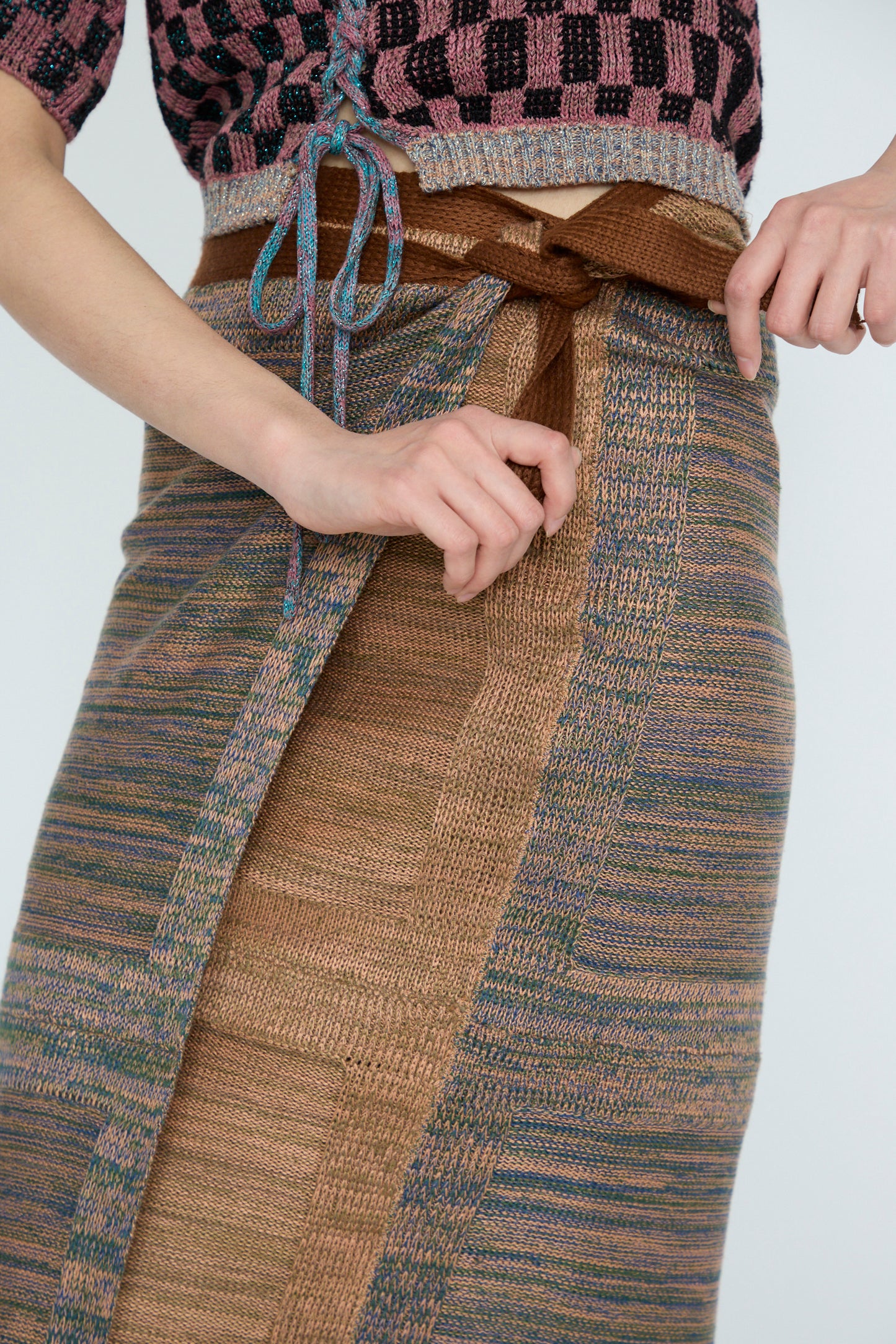 A person adjusts an Intensity Wrap Knitted Skirt in Multi with a brown waistband, against a white backdrop. 