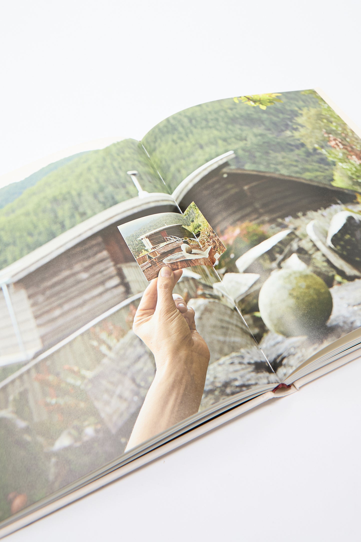 A hand holding the Fourth Edition, a small archival photograph of a cabin, against the backdrop of a similar cabin and garden displayed on an open book captures the essence of JB Blunk's artistic practice.