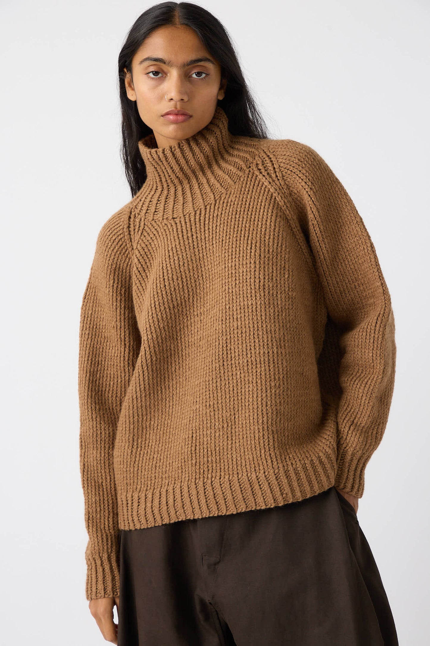 Person dressed in a Jan-Jan Van Essche Camel Wool Knit Sweater in Natural with a chunky turtleneck and dark pants stands against a plain background.