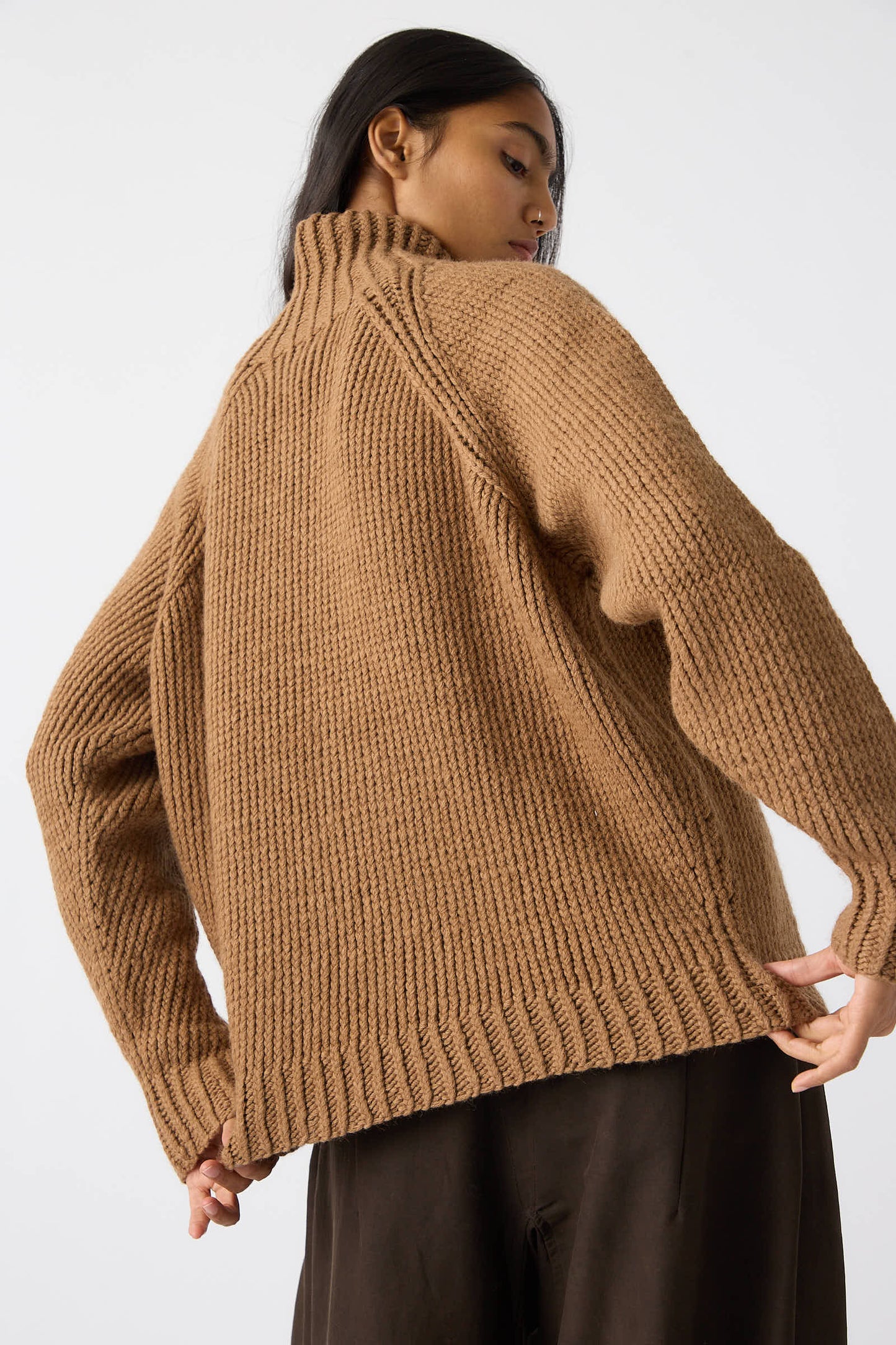 Person wearing a Jan-Jan Van Essche Camel Wool Knit Sweater in Natural, viewed from behind, with long black hair and dark-colored pants. The relaxed fit sweater is a rich brown color against the plain white background.