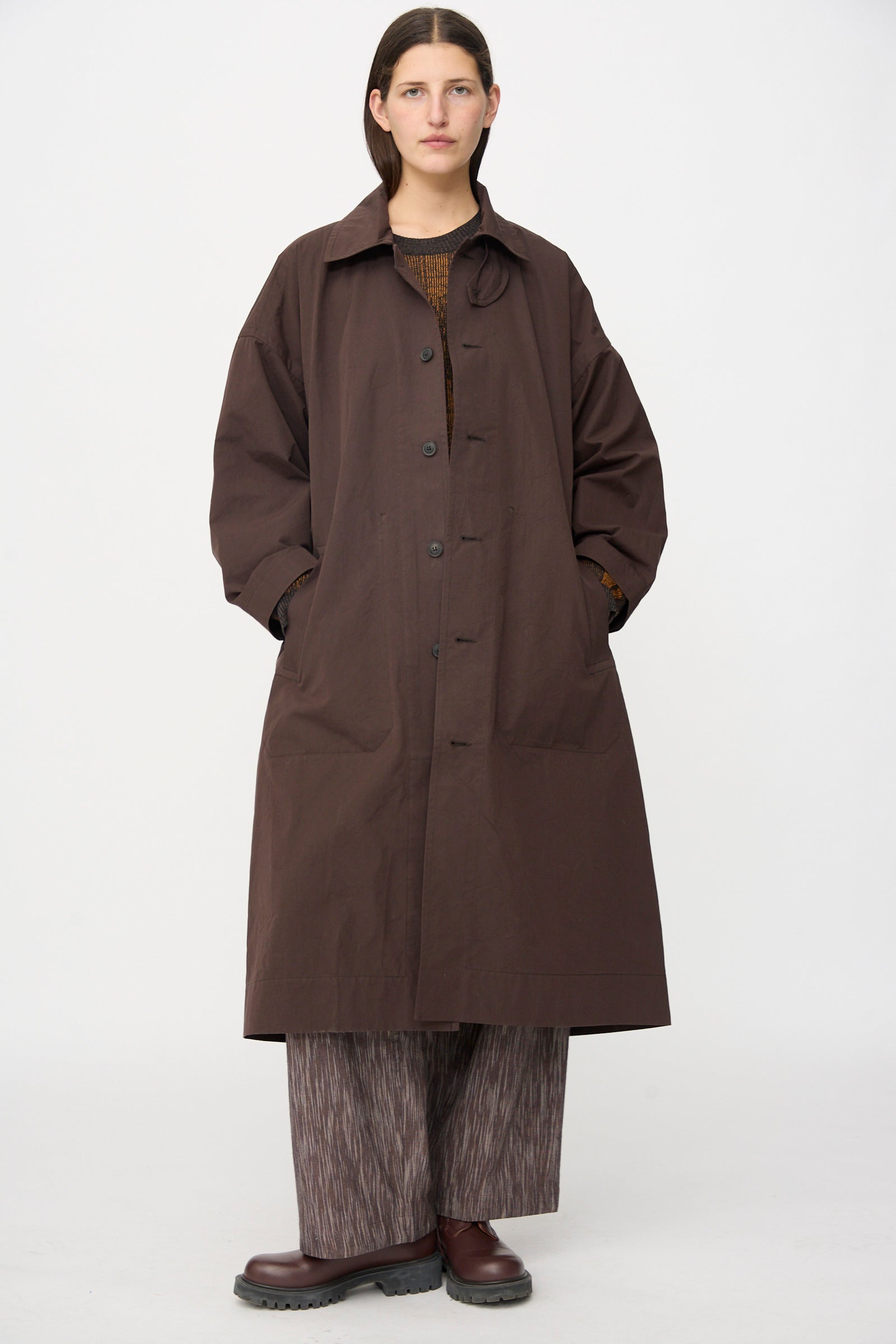 A person stands facing forward with hands in pockets, wearing the Jan-Jan Van Essche Cotton Silk Canvas Coat #34 in Old Mahogany, paired with patterned wide-leg pants and brown shoes against a plain white background.