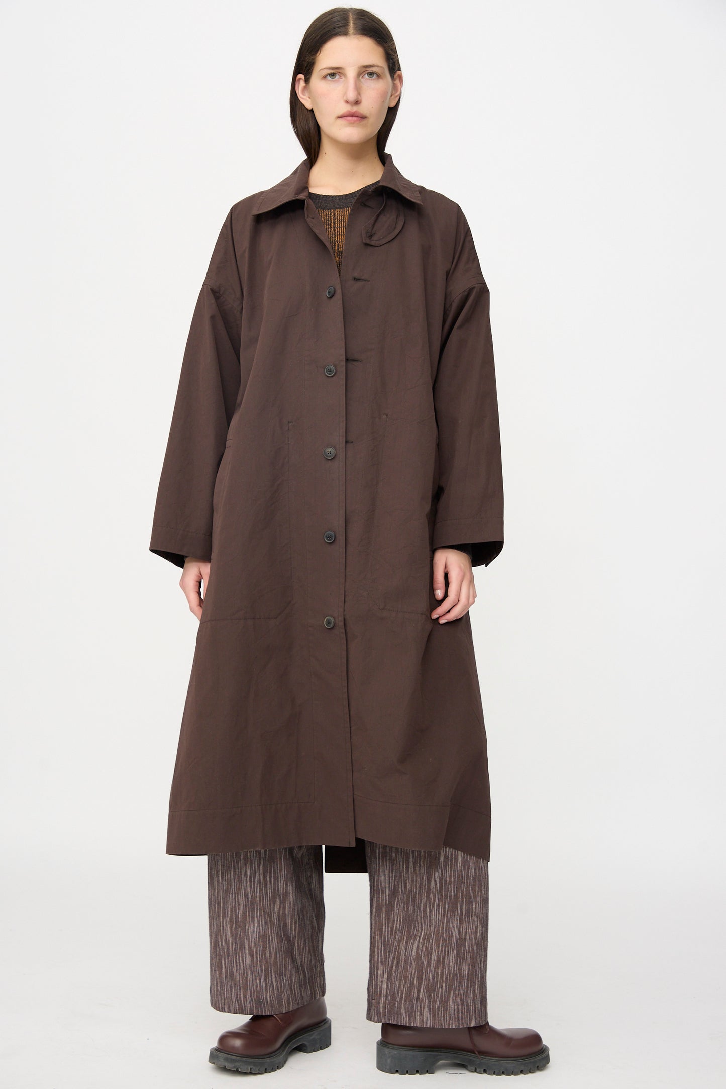 A person stands against a plain background, wearing the Jan-Jan Van Essche Cotton Silk Canvas Coat #34 in Old Mahogany, paired with patterned wide-leg pants and brown shoes.