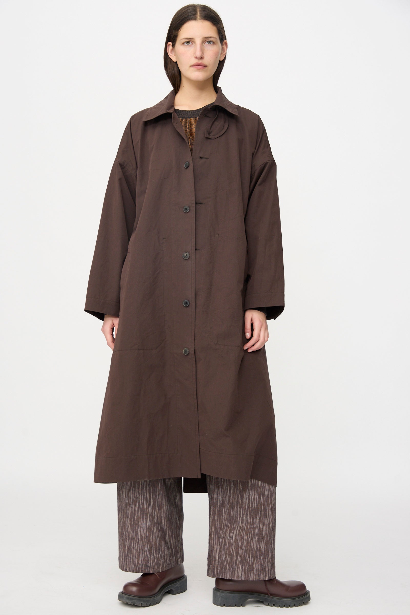 A person stands against a plain background, wearing the Jan-Jan Van Essche Cotton Silk Canvas Coat #34 in Old Mahogany, paired with patterned wide-leg pants and brown shoes.