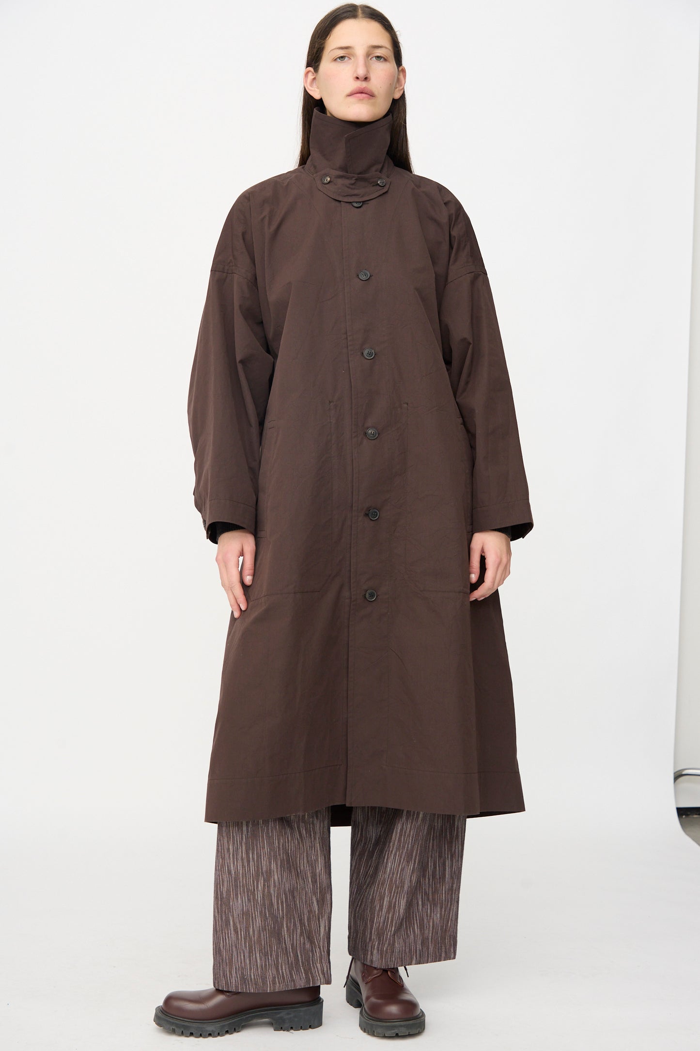 A person stands against a plain background wearing the Jan-Jan Van Essche Cotton Silk Canvas Coat #34 in Old Mahogany, paired with wide-leg patterned pants and brown shoes.