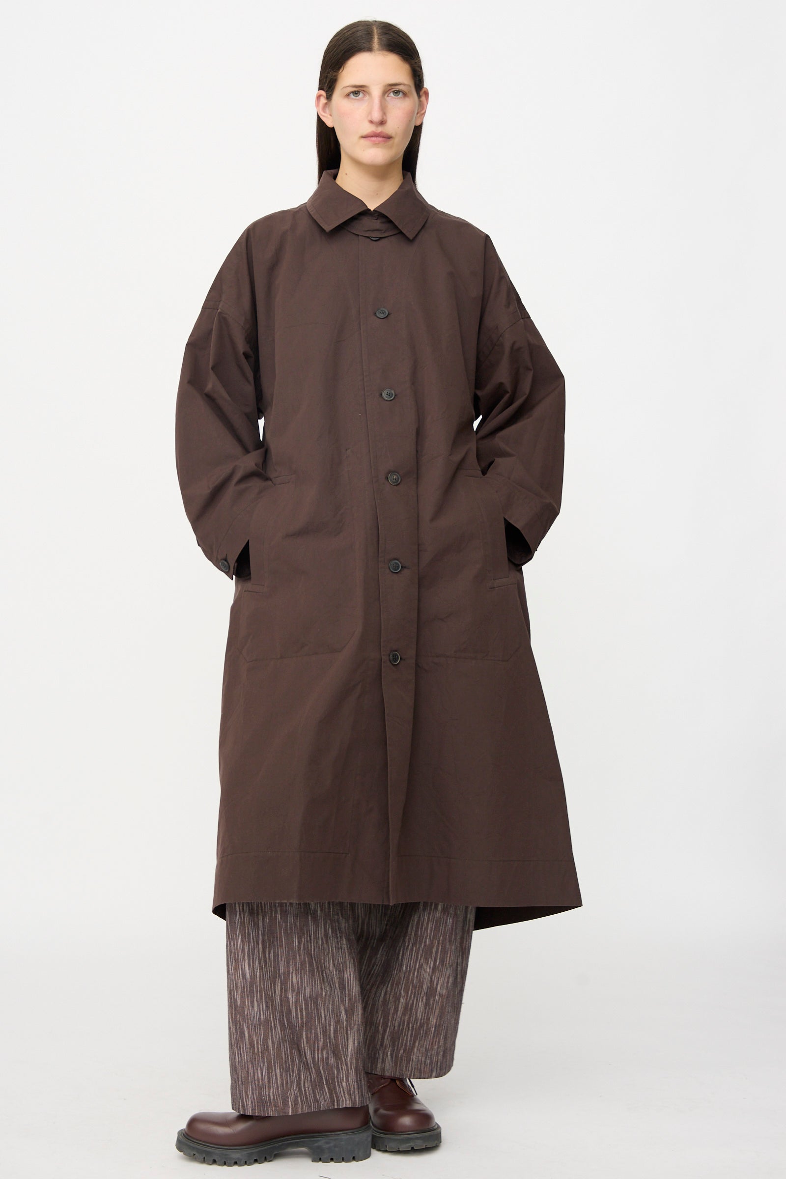 A person stands facing forward against a plain light background, wearing the Jan-Jan Van Essche Cotton Silk Canvas Coat #34 in Old Mahogany, paired with wide patterned pants and brown shoes.