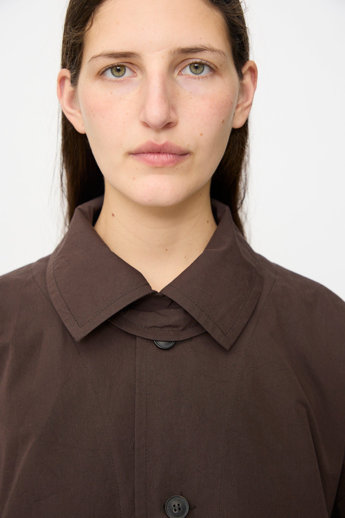 A person with long brown hair, neutral expression, and light skin wears the Jan-Jan Van Essche Cotton Silk Canvas Coat #34 in Old Mahogany, styled with a self-tie belt and photographed against a white background.