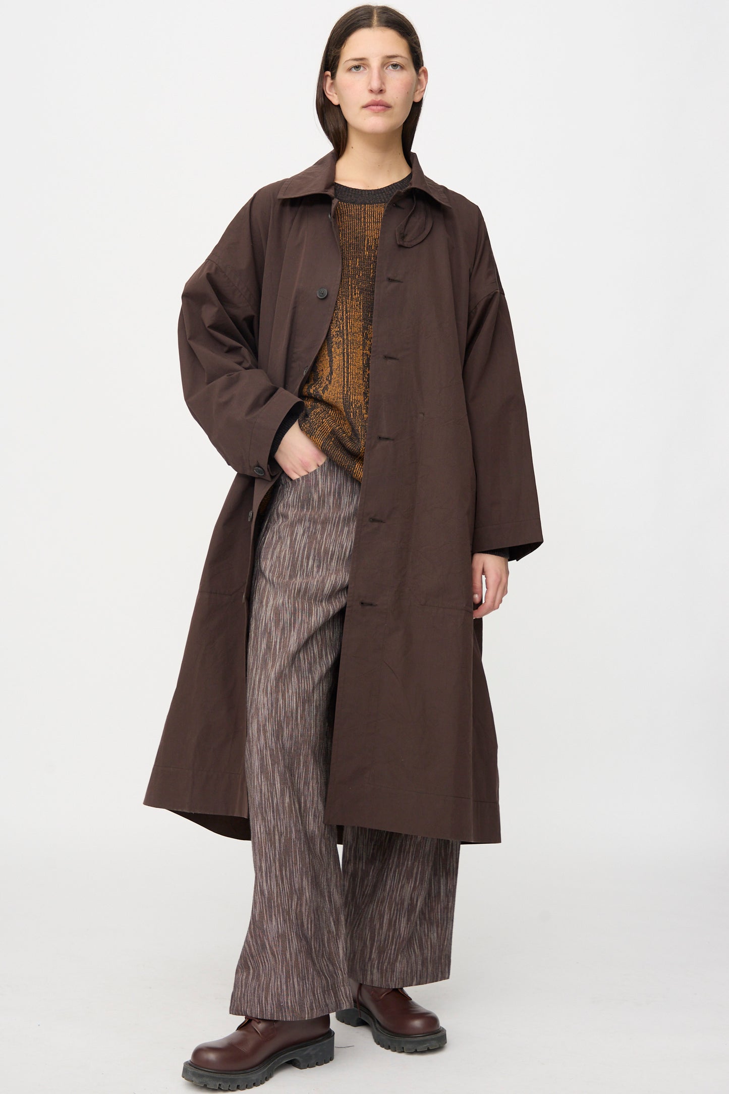 A person stands against a white background wearing the Jan-Jan Van Essche Cotton Silk Canvas Coat #34 in Old Mahogany, paired with a patterned brown sweater, wide-legged striped pants, and brown shoes.