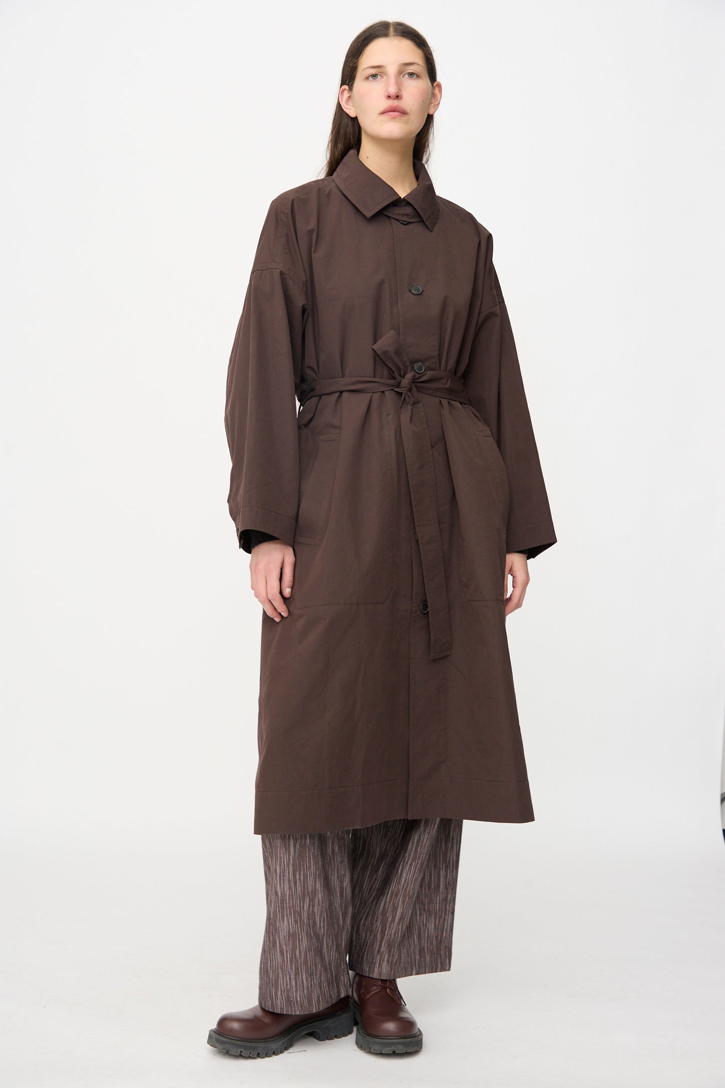 A person stands against a plain background wearing the Jan-Jan Van Essche Cotton Silk Canvas Coat #34 in Old Mahogany, styled with loose patterned pants and brown shoes.