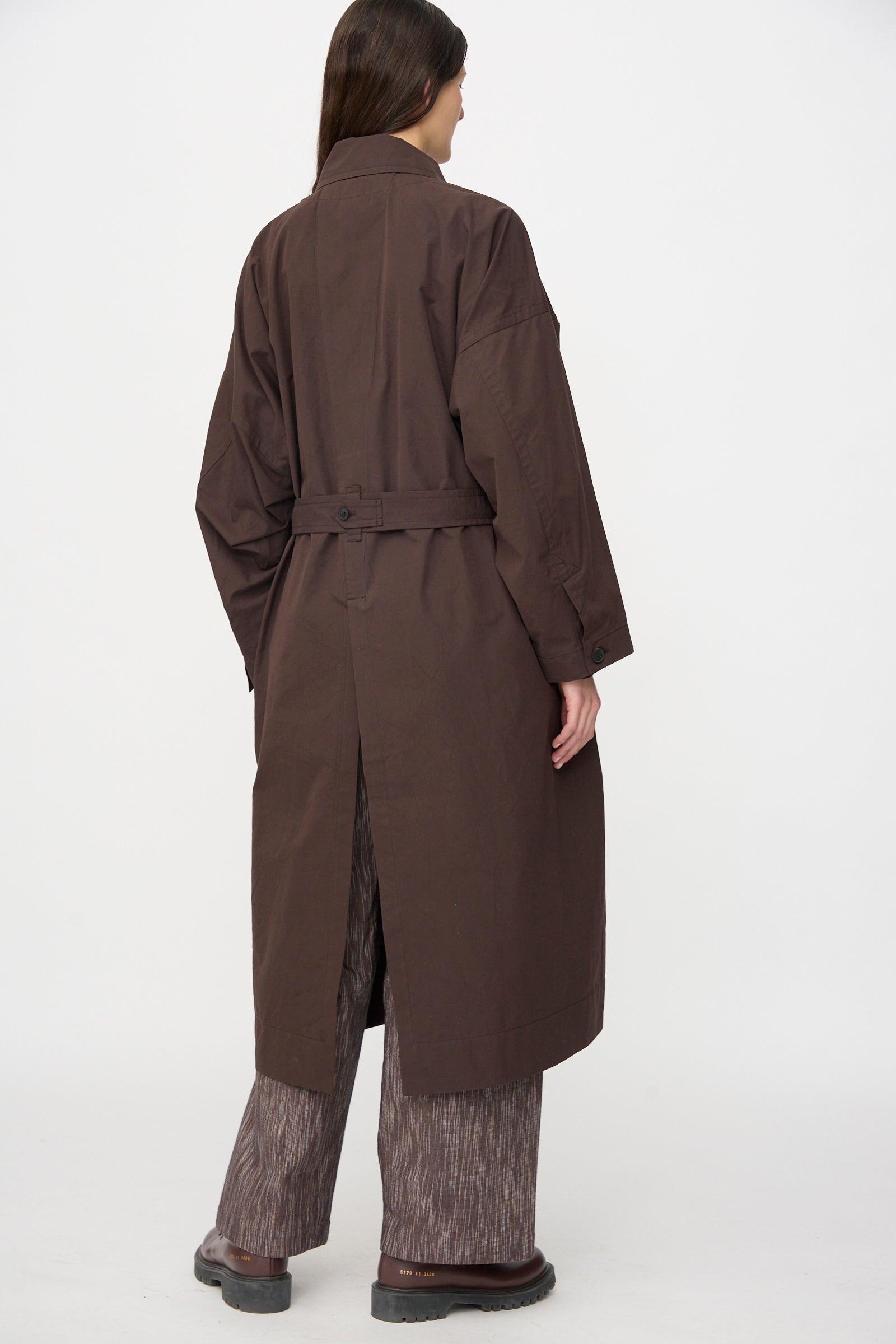 A person stands facing away, wearing the Jan-Jan Van Essche Cotton Silk Canvas Coat #34 in Old Mahogany, styled with wide-legged patterned pants against a plain white background.
