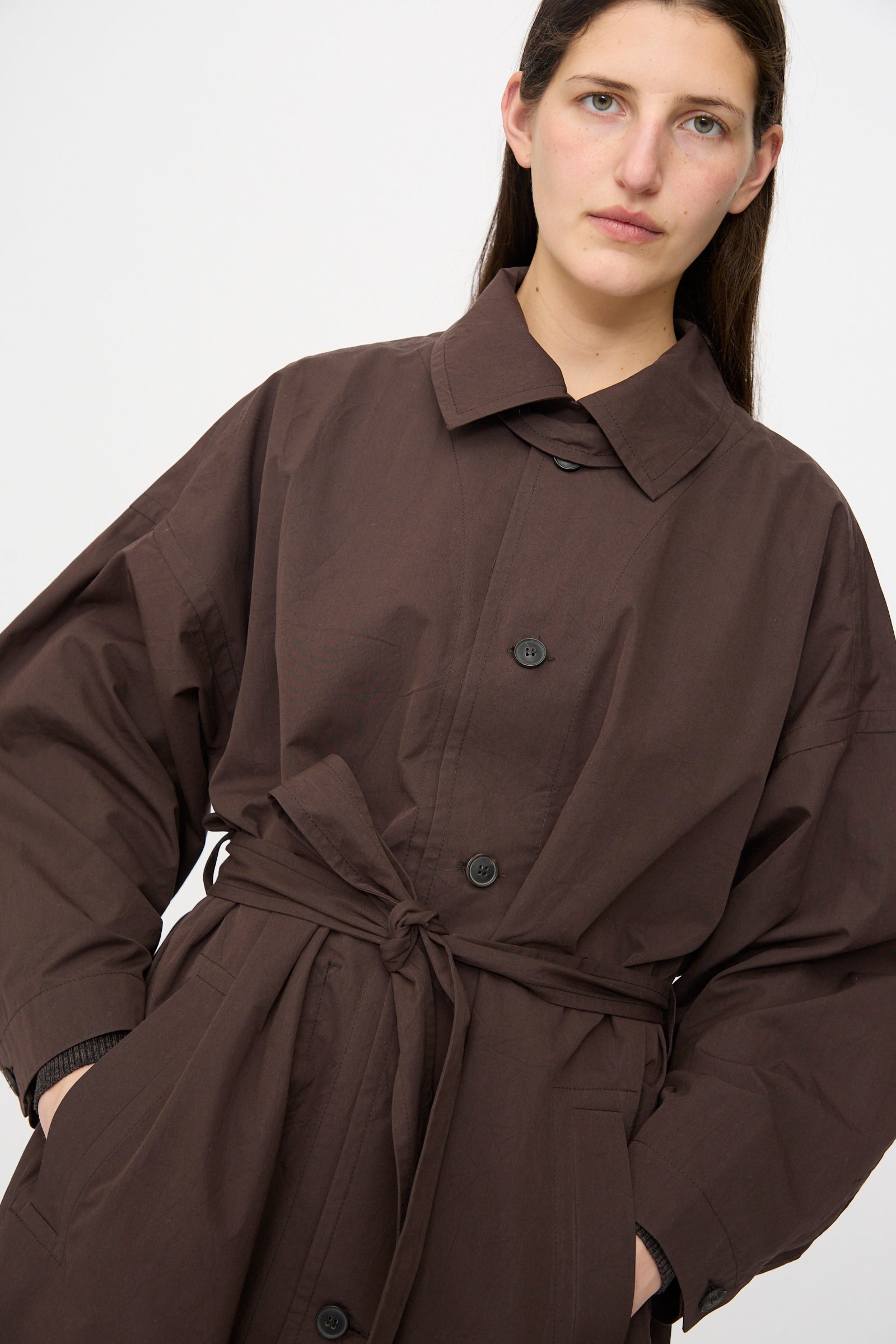 A person with long brown hair wears the Jan-Jan Van Essche Cotton Silk Canvas Coat #34 in Old Mahogany, featuring a loose fit and self-tie belt, standing against a plain white background.
