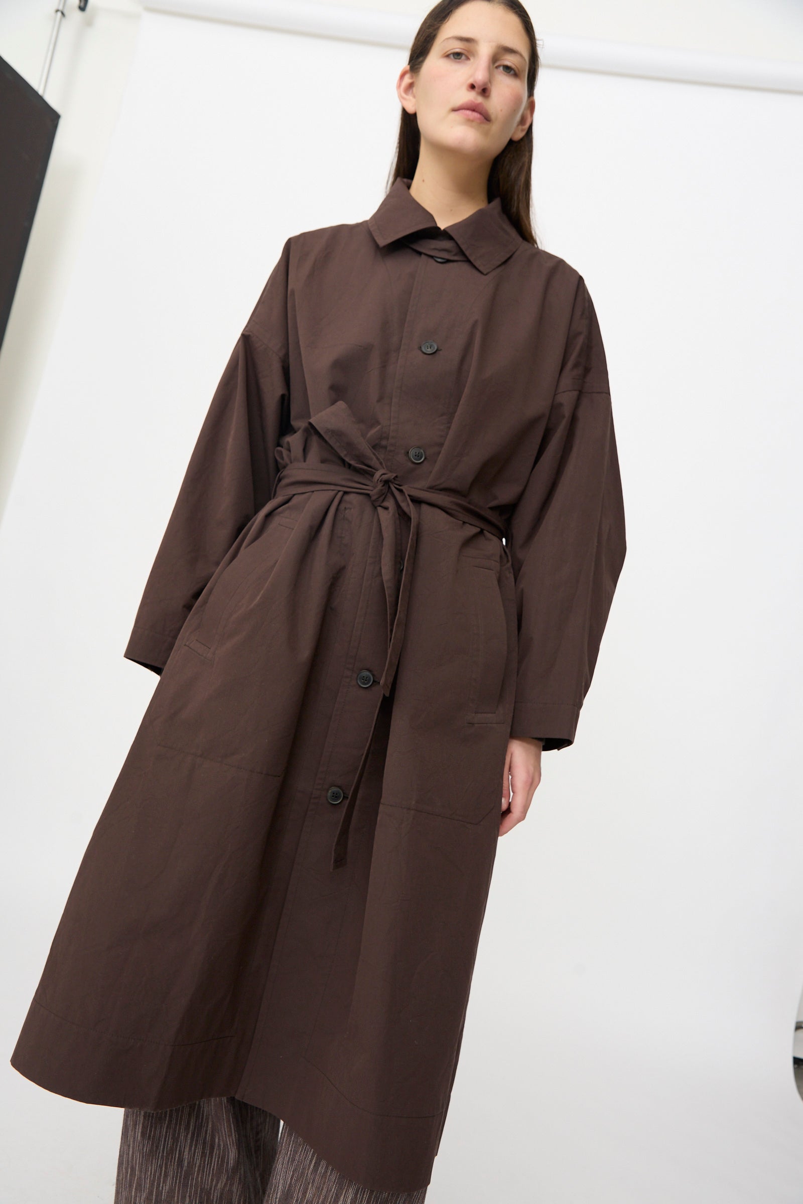 A person stands against a plain white background wearing the Jan-Jan Van Essche Cotton Silk Canvas Coat #34 in Old Mahogany, featuring loose sleeves and a self-tie belt at the waist.