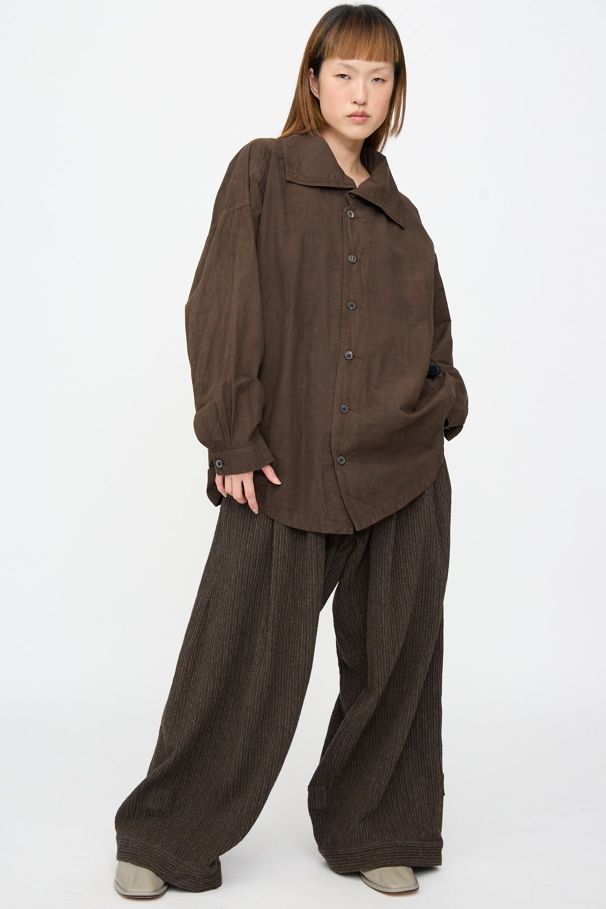 A person wearing a Cotton Wool Shirt #110 in Doro Zome, an oversized dark brown jacket, with wide-leg pinstriped trousers.
