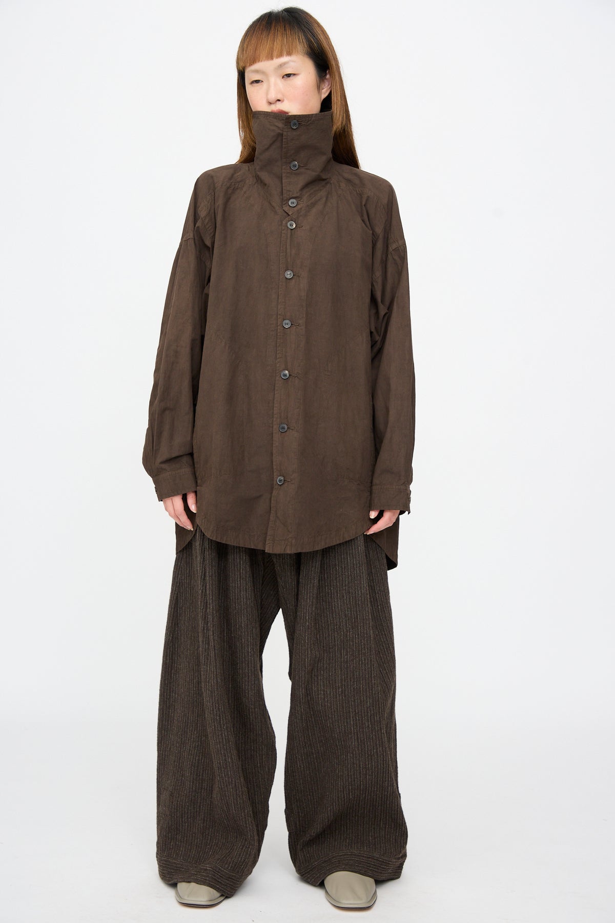 A person wears a Cotton Wool Shirt #110 in Doro Zome over an oversized shirt jacket with a high collar and wide-legged, dark brown striped trousers.
