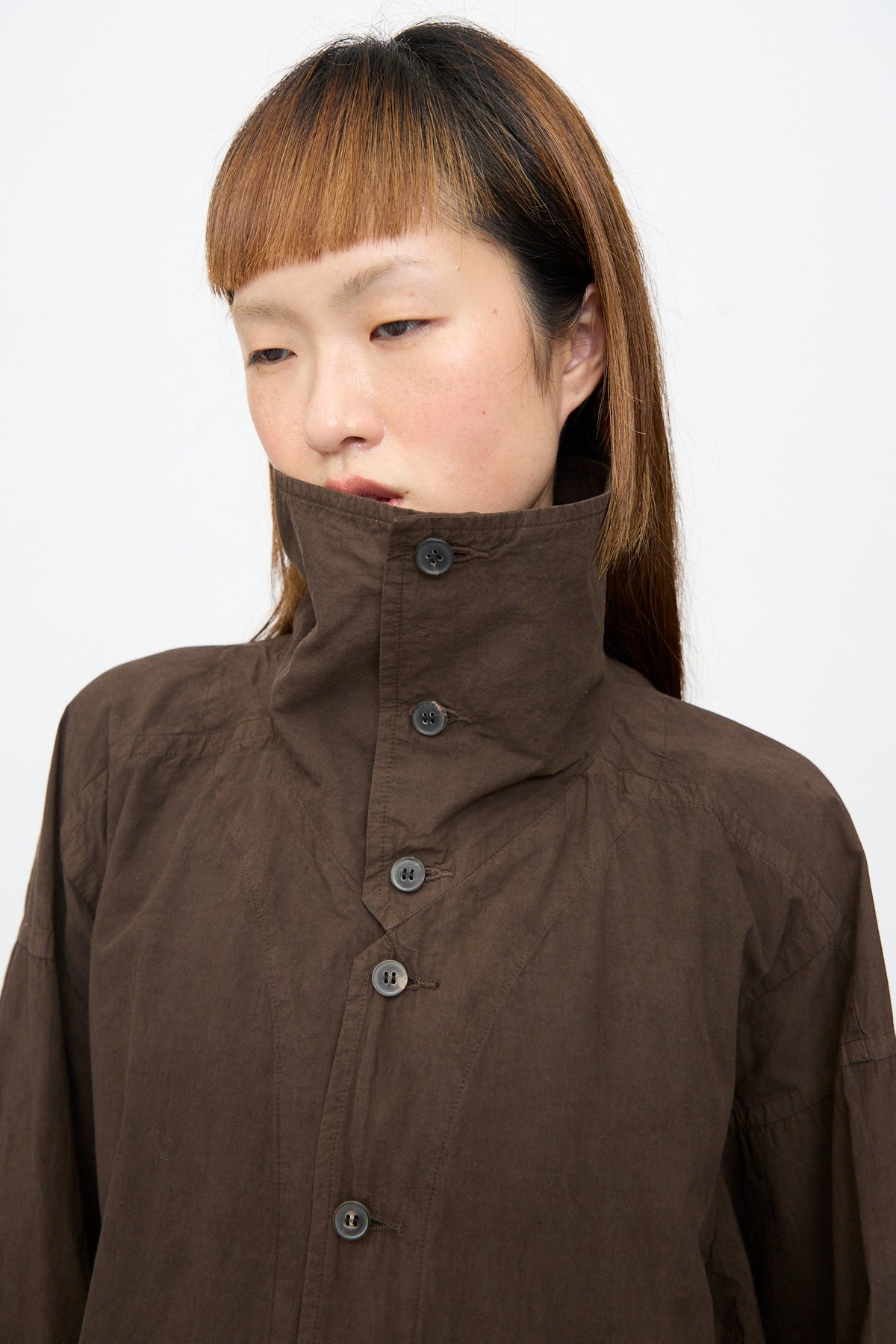 A person wearing a dark brown Cotton Wool Shirt #110 in Doro Zome with an oversized fit and a high collar covering their lower face.