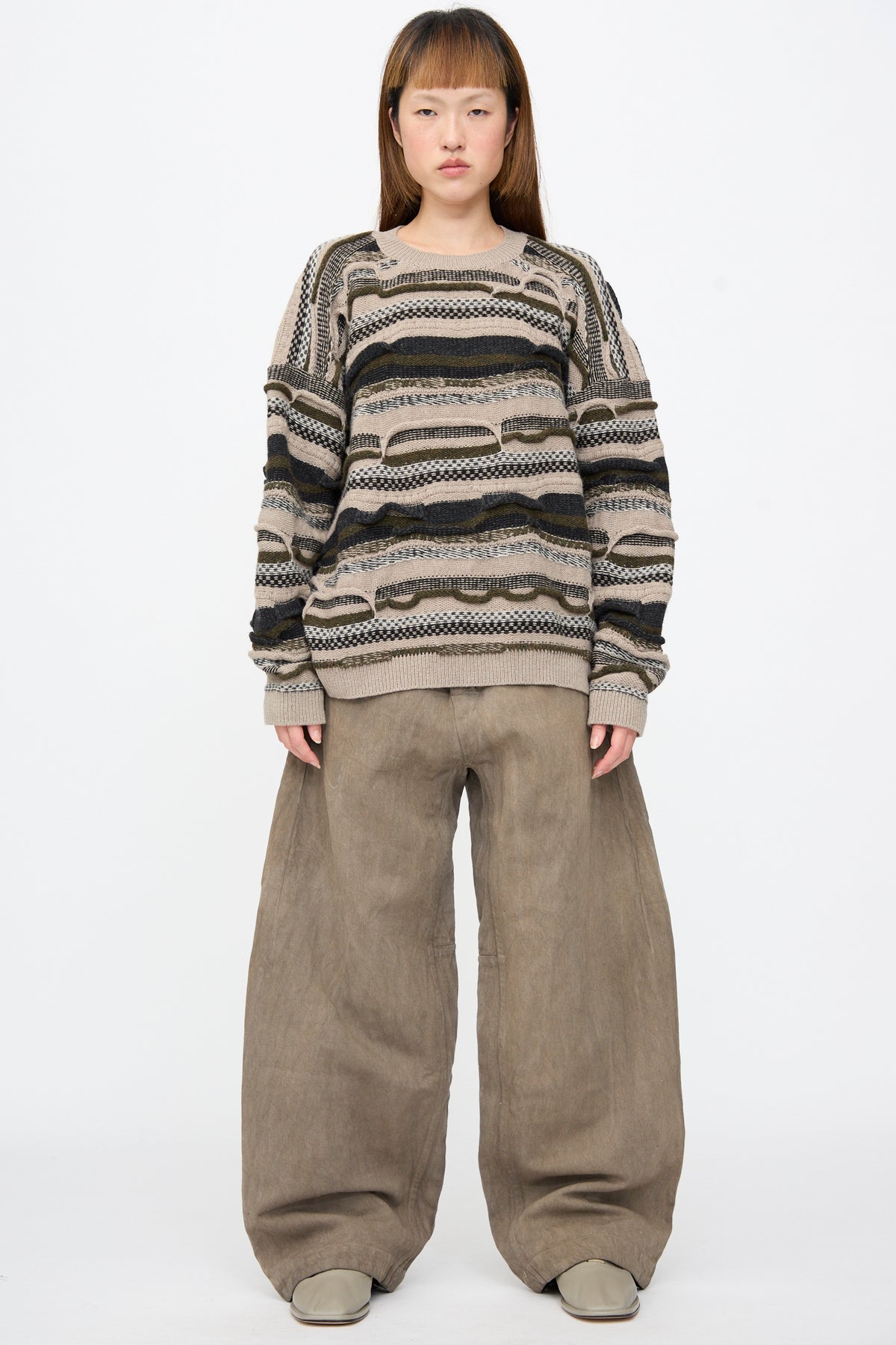 A person wears a long-sleeved, horizontally striped, textured sweater in beige, olive, and black, paired with Jan-Jan Van Essche Hemp Bamboo Twill Trousers #90 in Kakishibu and Iron.