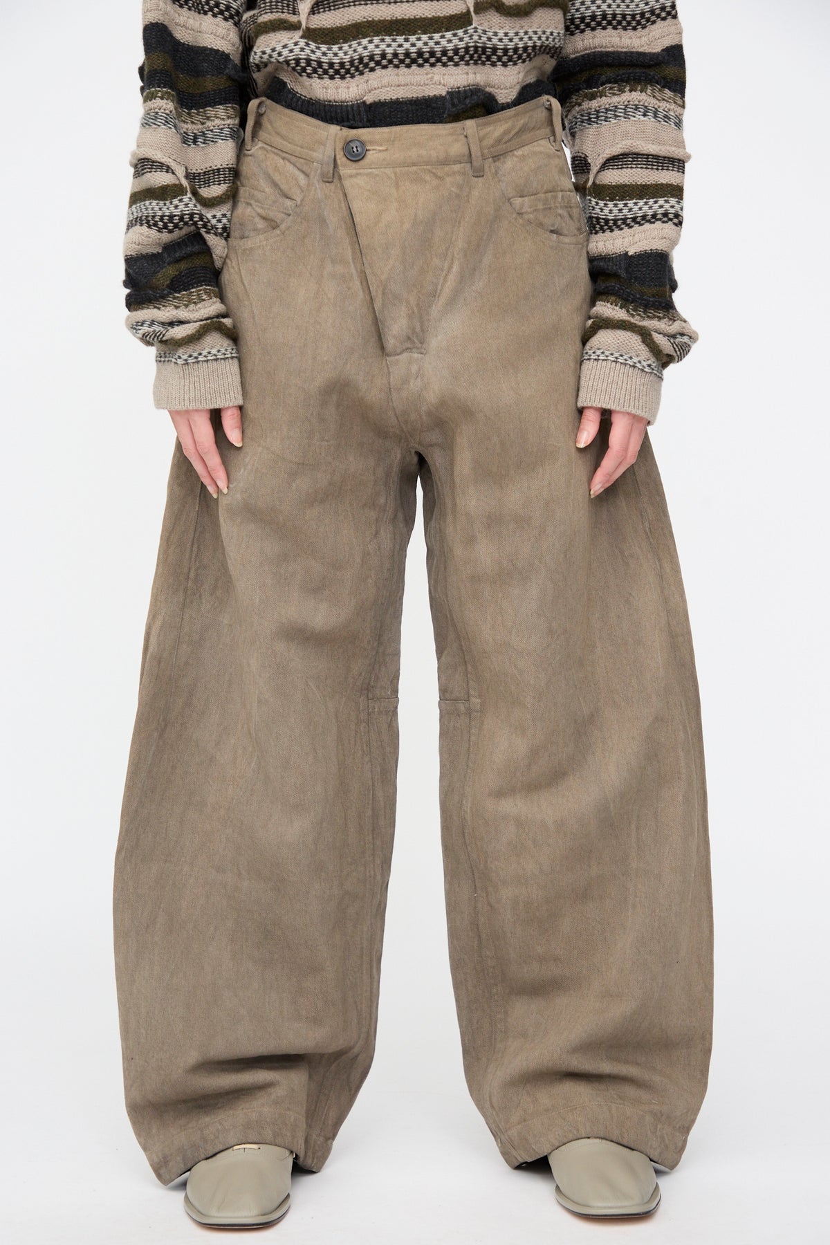 A person wearing Hemp Bamboo Twill Trousers #90 in Kakishibu and Iron, featuring wide-leg, muted brown fabric with an asymmetrical overlapping button closure, paired with a striped sweater and light shoes.