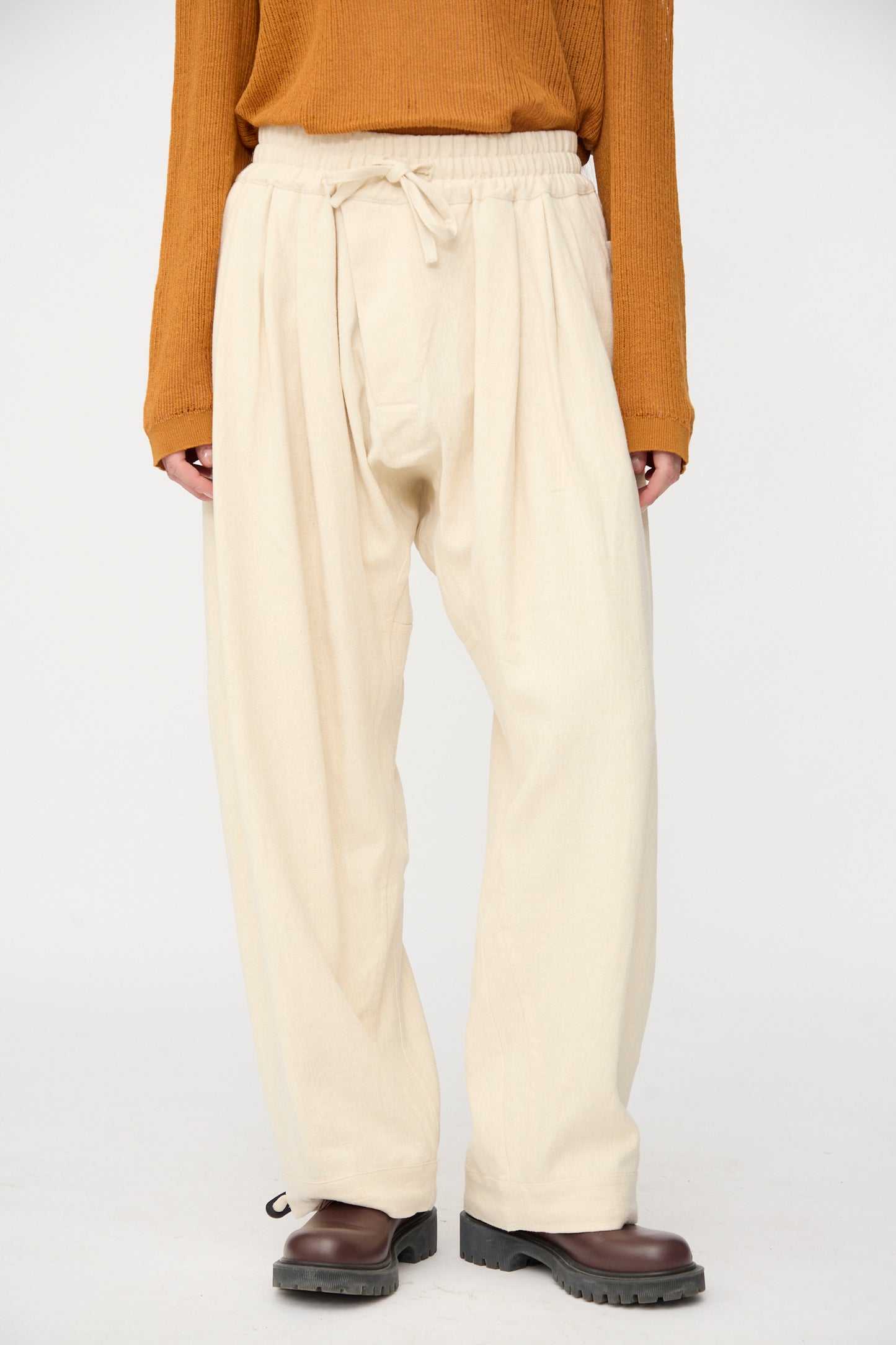 A person wearing Jan-Jan Van Essche Hemp Soft Canvas Trousers #91 in Kinari with an elasticated waistband, a rust long sleeve top, and dark brown shoes stands against a plain light background.