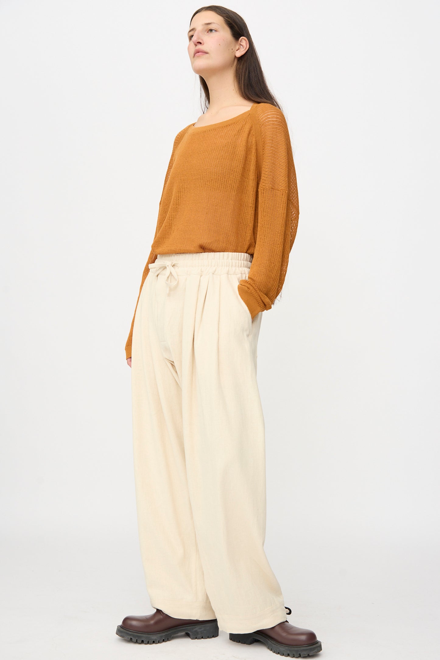 A person stands against a plain background wearing a loose fit brown long-sleeve top, Jan-Jan Van Essche Hemp Soft Canvas Trousers #91 in Kinari with an elasticated waistband, and dark brown shoes.