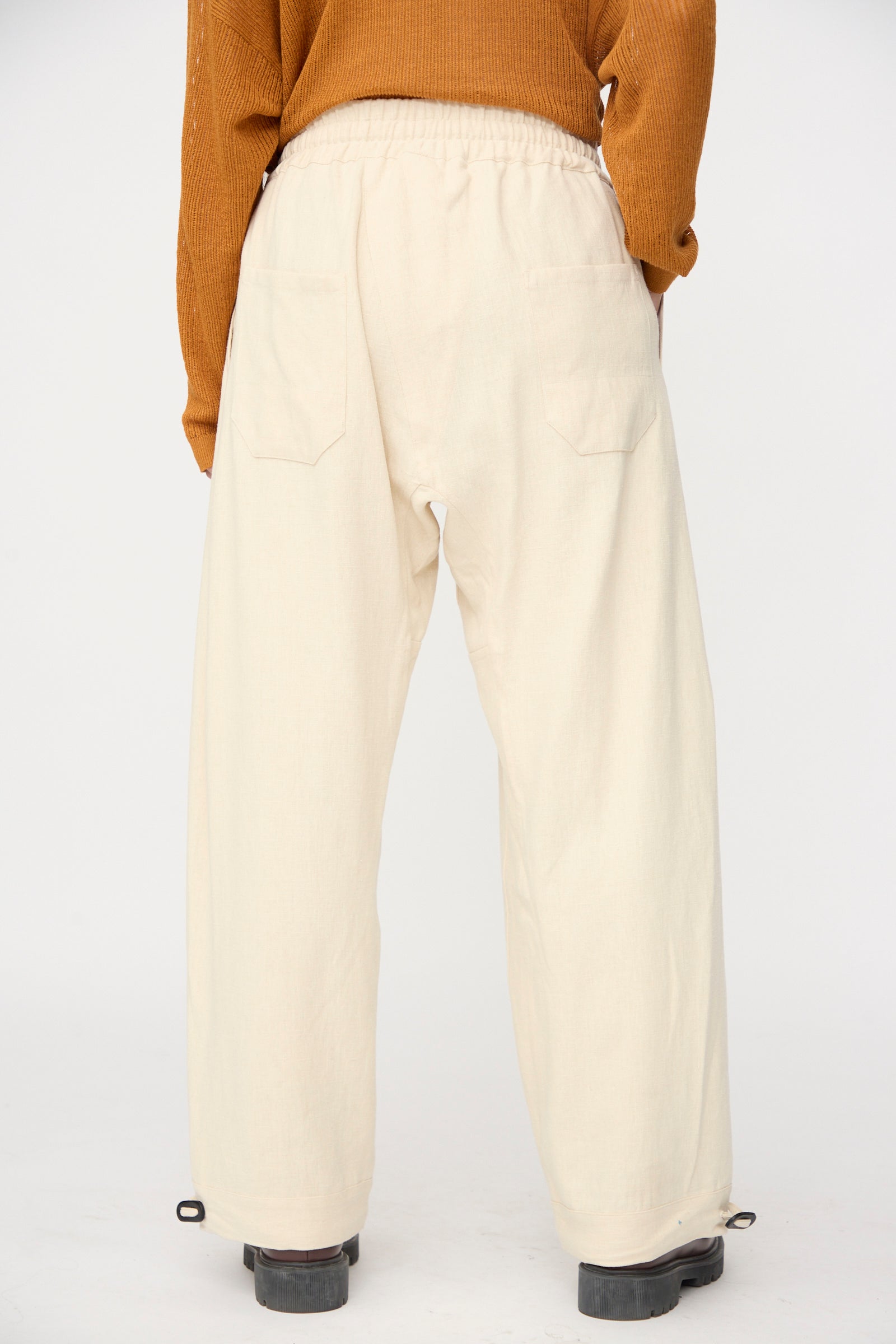 A person stands with their back to the camera in Jan-Jan Van Essche Hemp Soft Canvas Trousers #91 in Kinari, featuring a loose fit and elasticated waistband, paired with a brown sweater against a plain background.