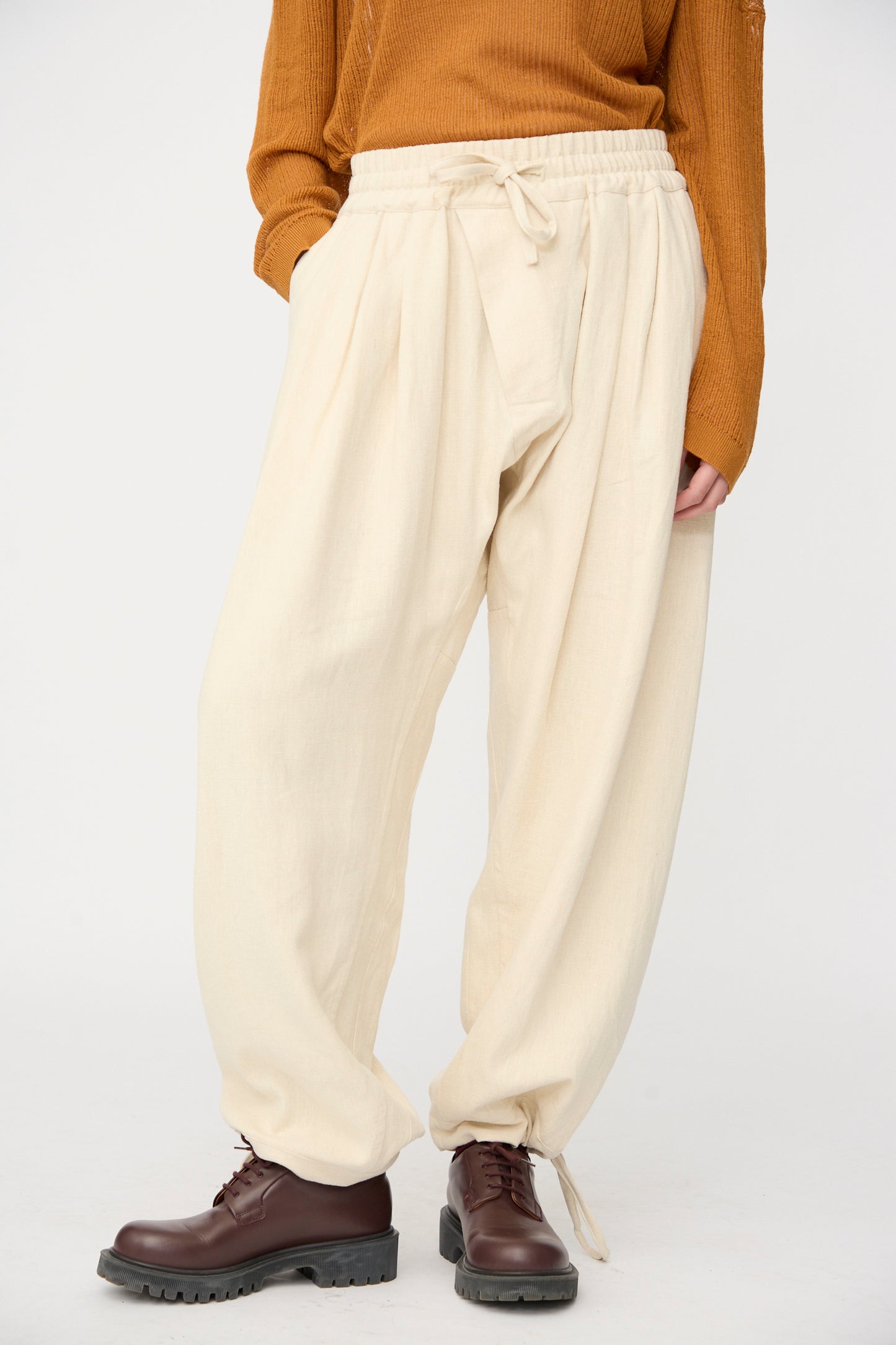 A person wears Jan-Jan Van Essche Hemp Soft Canvas Trousers #91 in Kinari—cream-colored, loose-fit with an elasticated waistband—paired with a brown sweater and dark brown lace-up boots against a plain light background.