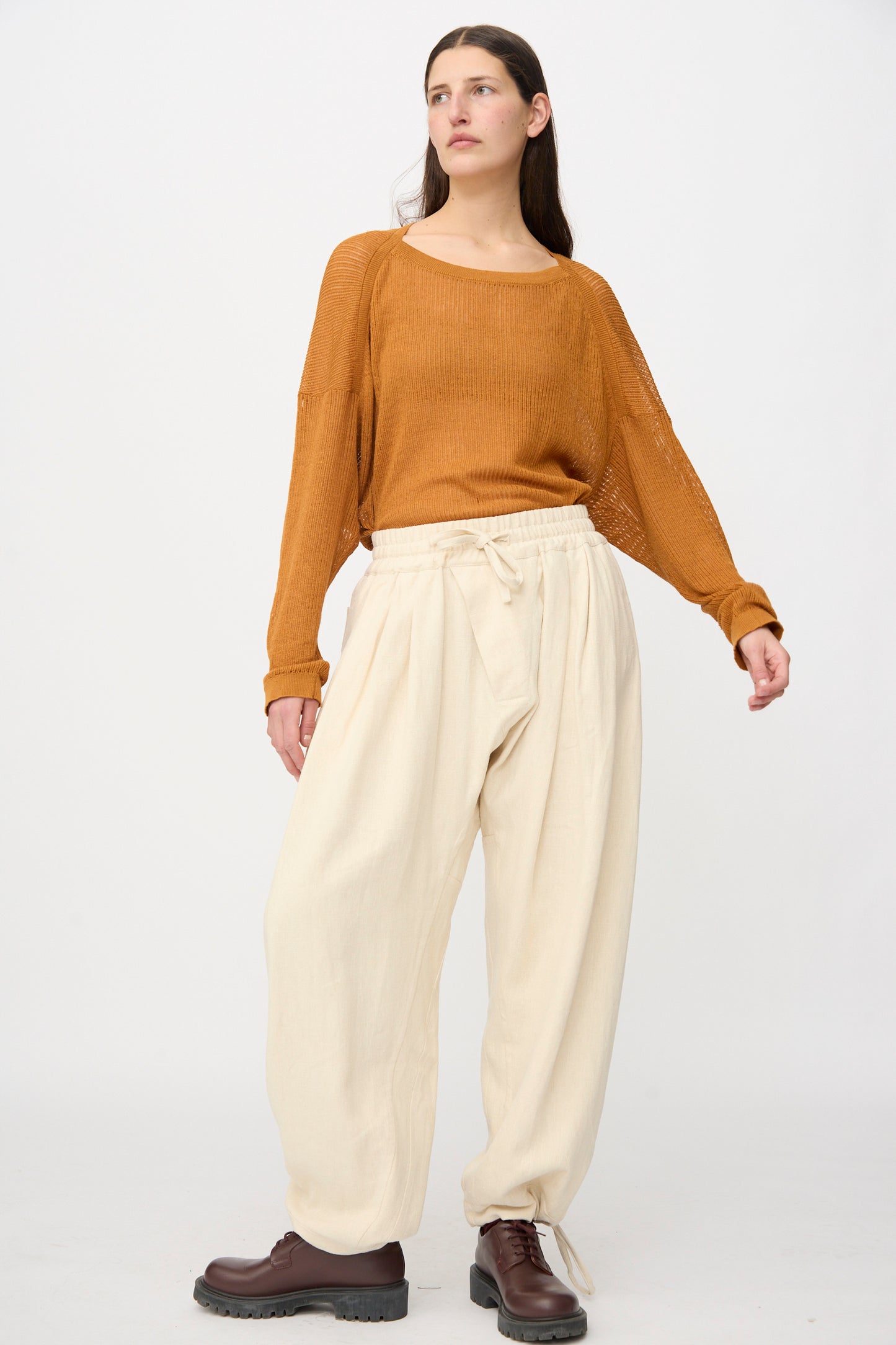 A person stands against a plain background wearing a loose brown sweater, brown lace-up boots, and Jan-Jan Van Essche Hemp Soft Canvas Trousers #91 in Kinari with an elasticated waistband.