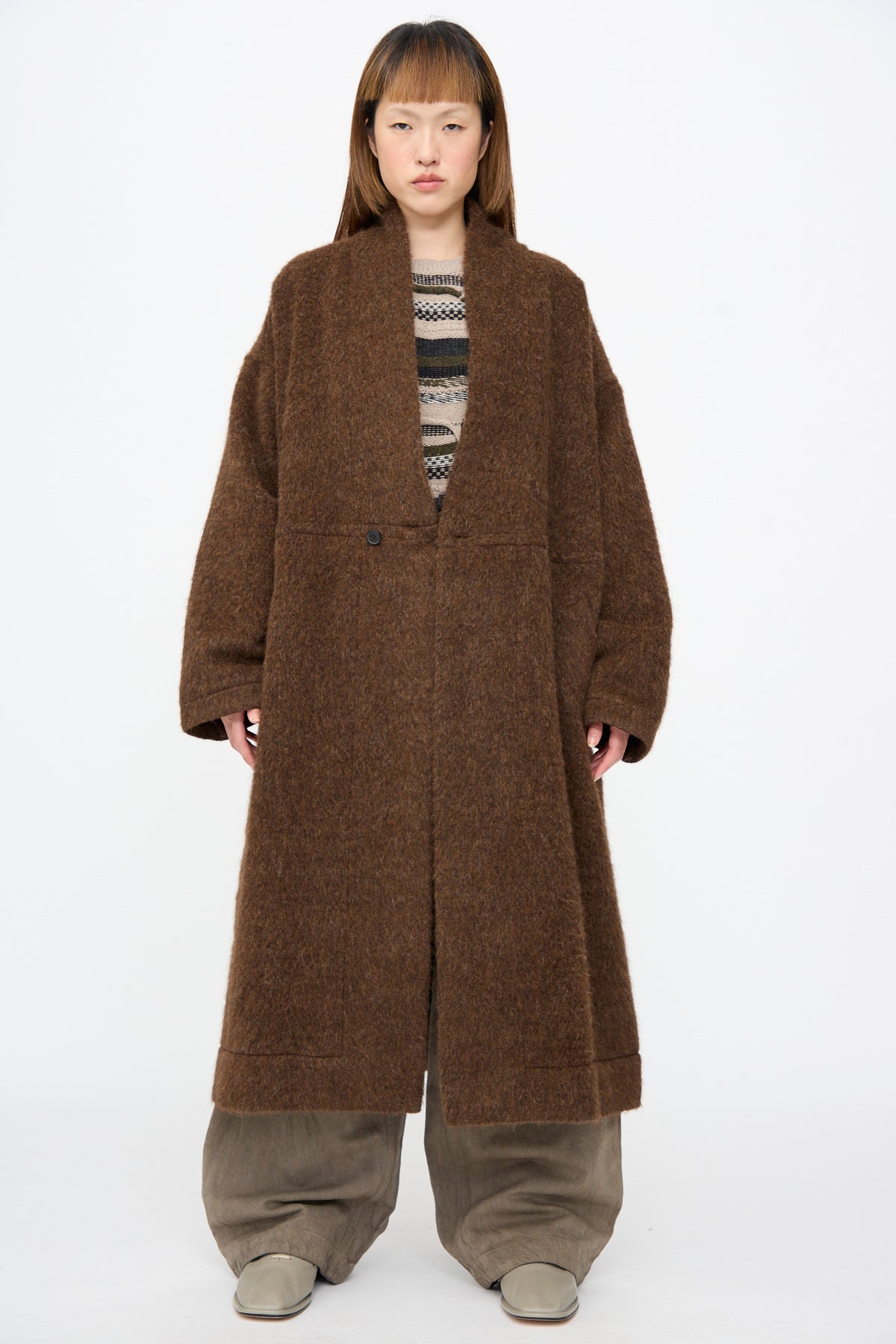 A person wearing a Jan-Jan Van Essche Highpile Herringbone Coat #35 in Brown Alpaca, styled with a single button closure over a patterned sweater and wide-leg trousers.