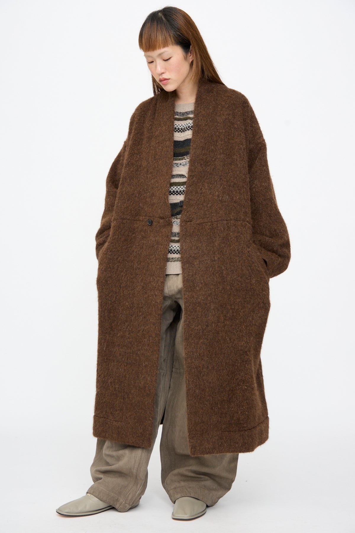 A person wearing a Jan-Jan Van Essche Highpile Herringbone Coat #35 in Brown Alpaca, an oversized single-button wool coat, a patterned sweater, and wide-leg trousers.