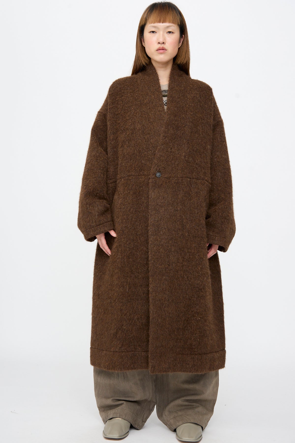 A person wearing a Jan-Jan Van Essche Highpile Herringbone Coat #35 in Brown Alpaca, an oversized wool coat with a single front button, over loose trousers and light-colored shoes.
