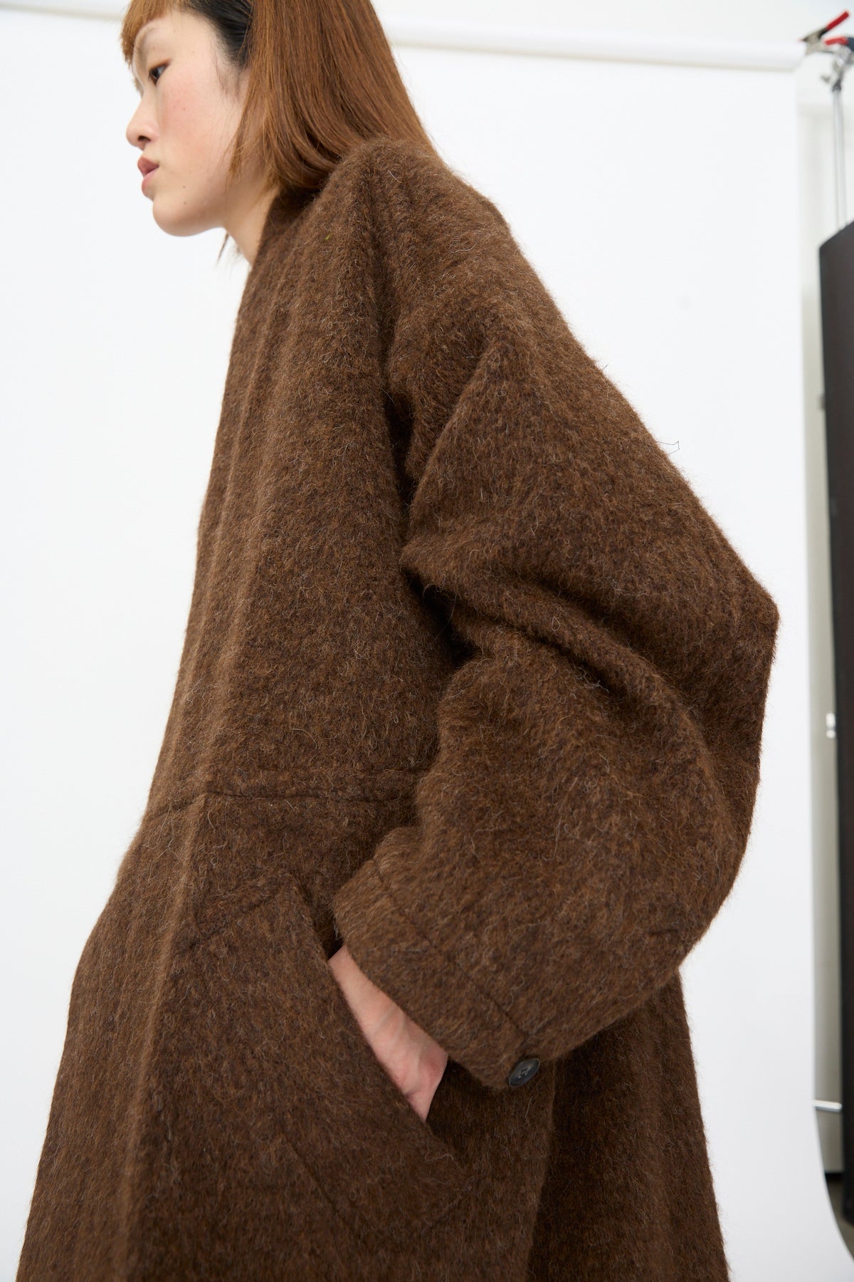 A person wearing a Jan-Jan Van Essche Highpile Herringbone Coat #35 in Brown Alpaca, an oversized brown wool coat with a textured, fuzzy finish and side pockets.