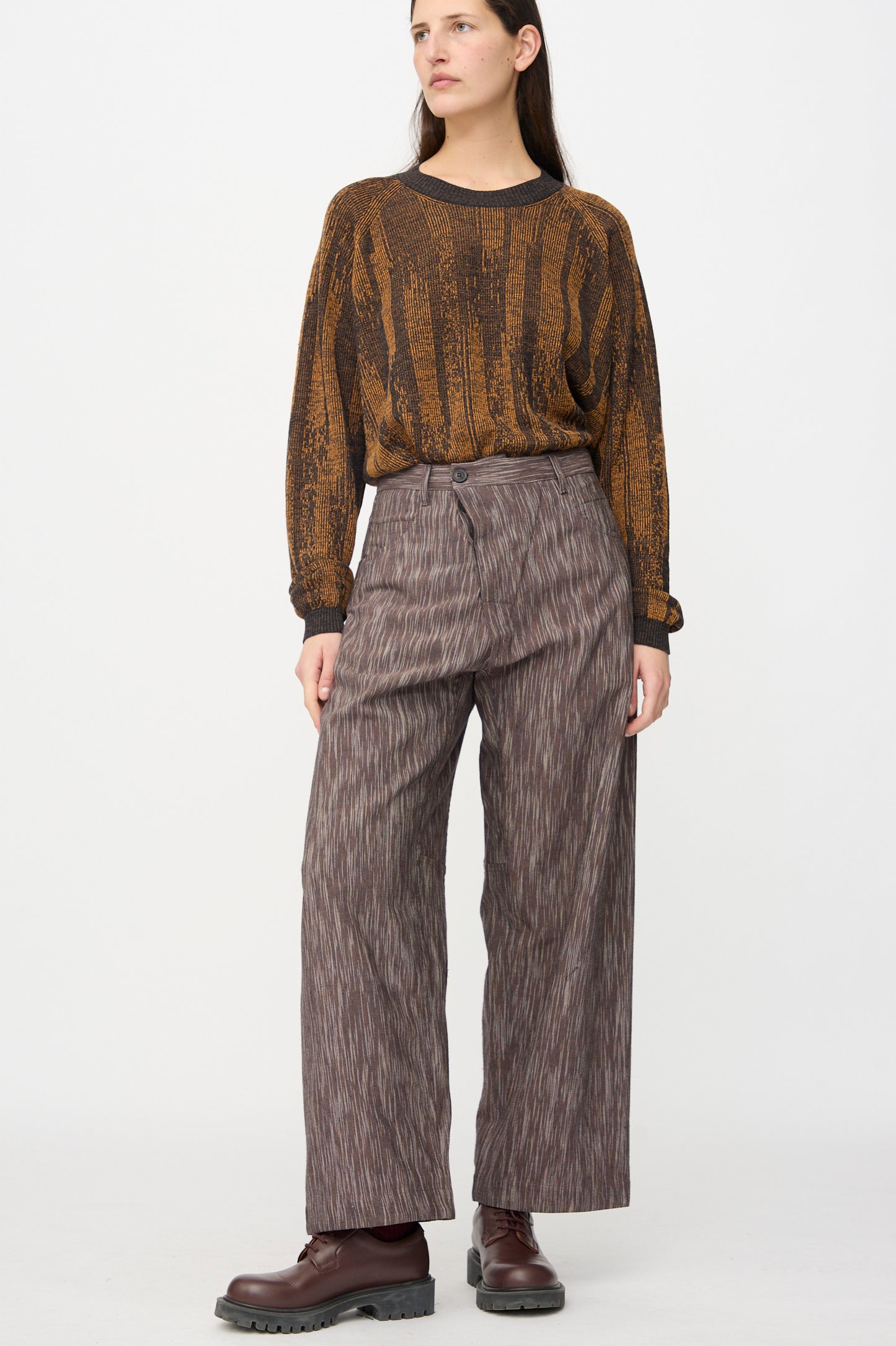A woman stands against a white background wearing Jan-Jan Van Essche Kasuri Cotton Silk Trousers #90 in Old Lavender, a brown patterned long-sleeved shirt, and brown boots.