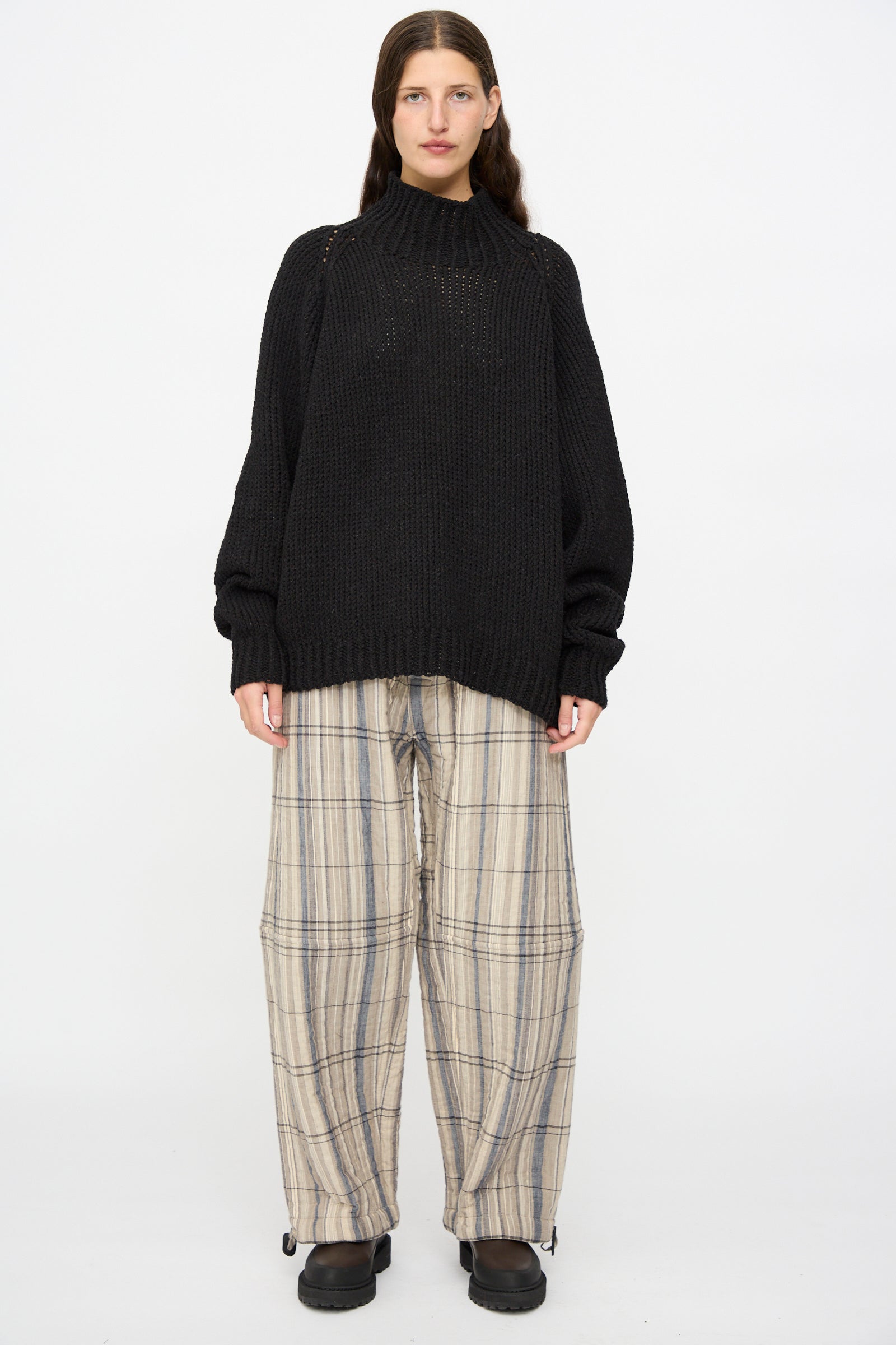A person stands facing forward wearing the Jan-Jan Van Essche Lalin Knit #68 Sweater in Pitch Black, paired with wide beige plaid pants and black shoes, set against a plain white background.