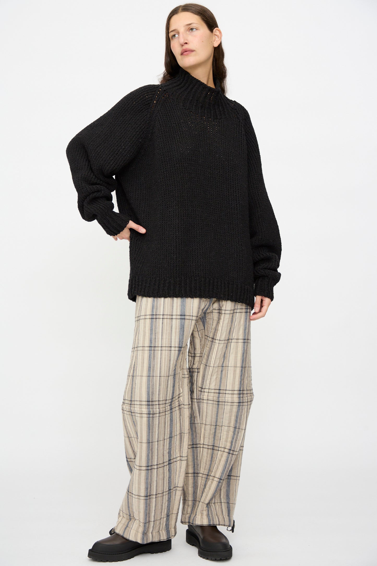 A person stands against a plain background wearing the Jan-Jan Van Essche Lalin Knit #68 Sweater in Pitch Black, paired with wide-leg beige plaid pants and black shoes.