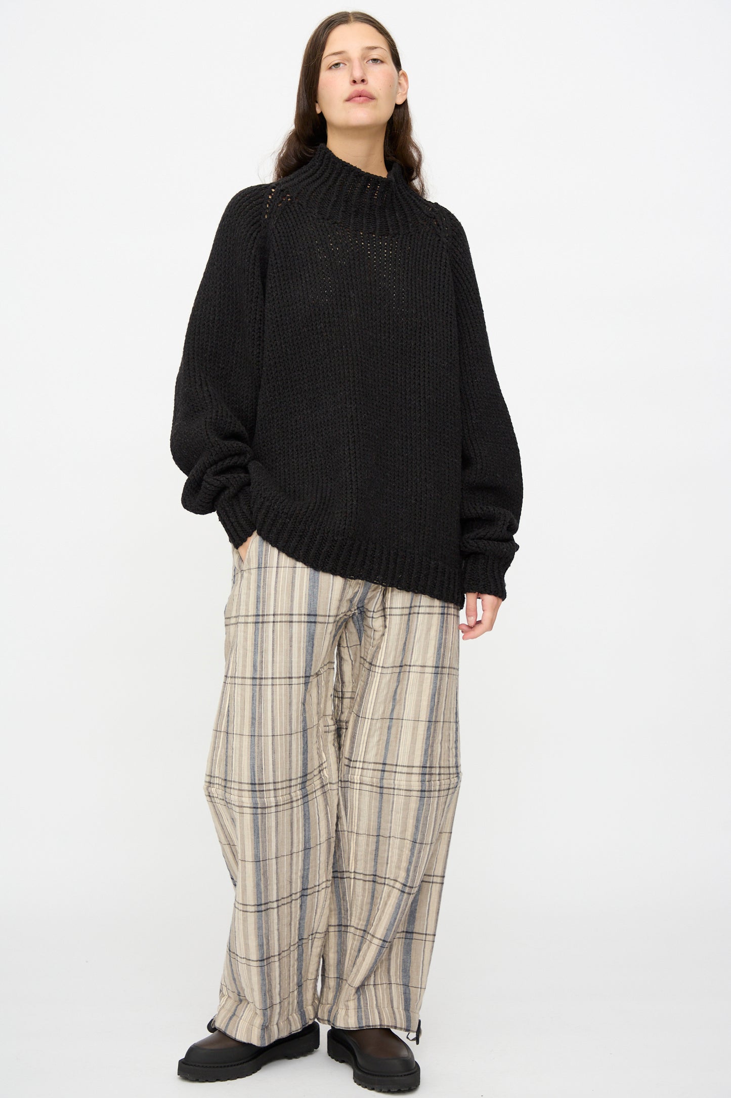 Person stands against a plain background wearing the Jan-Jan Van Essche Lalin Knit #68 Sweater in Pitch Black, paired with wide-legged plaid pants and black shoes.