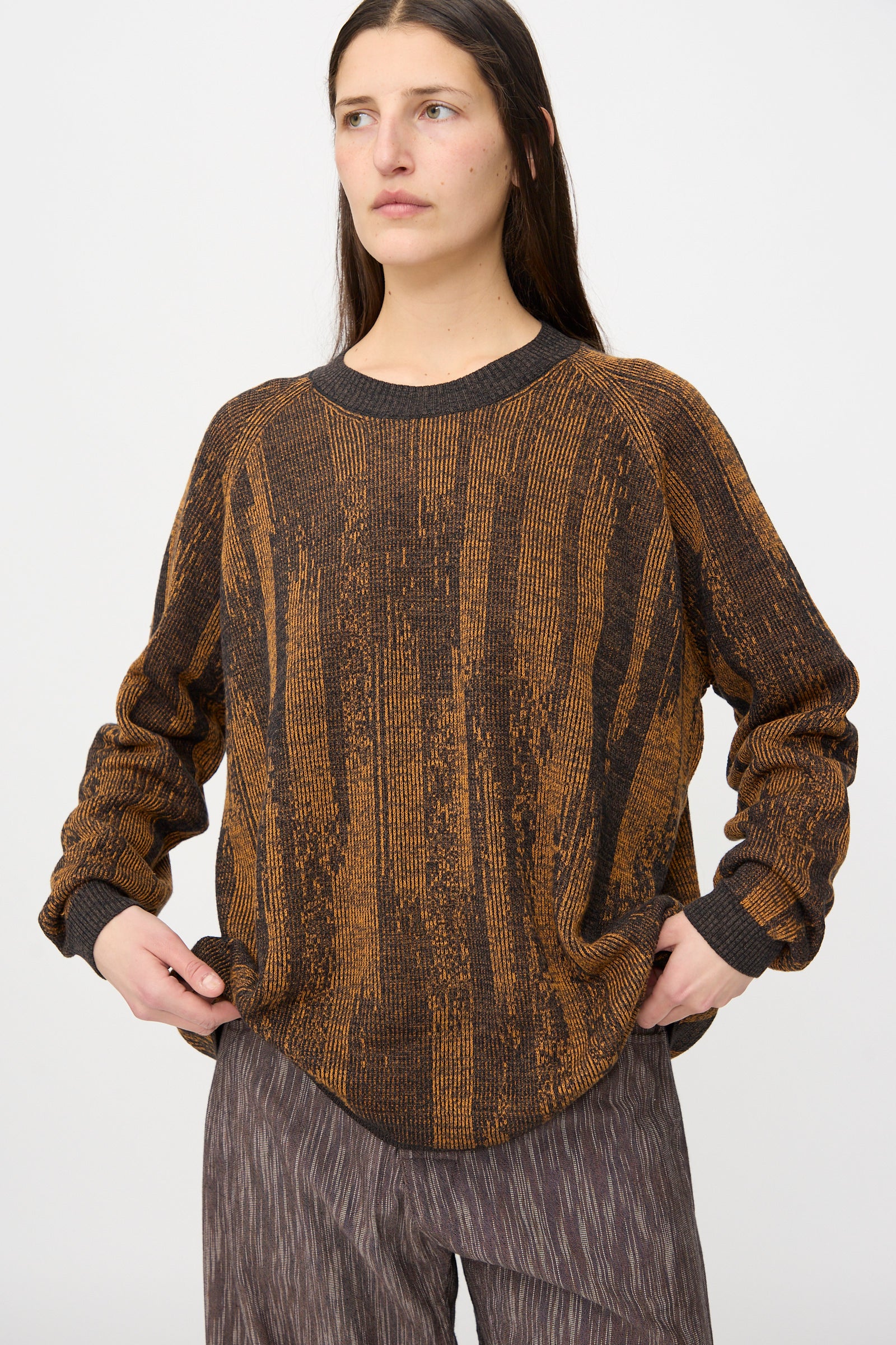 A woman stands against a plain white background wearing the Jan-Jan Van Essche Linen Cotton Kasuri Knit #77 in Black Brown Orange with long sleeves, paired with brown textured pants.