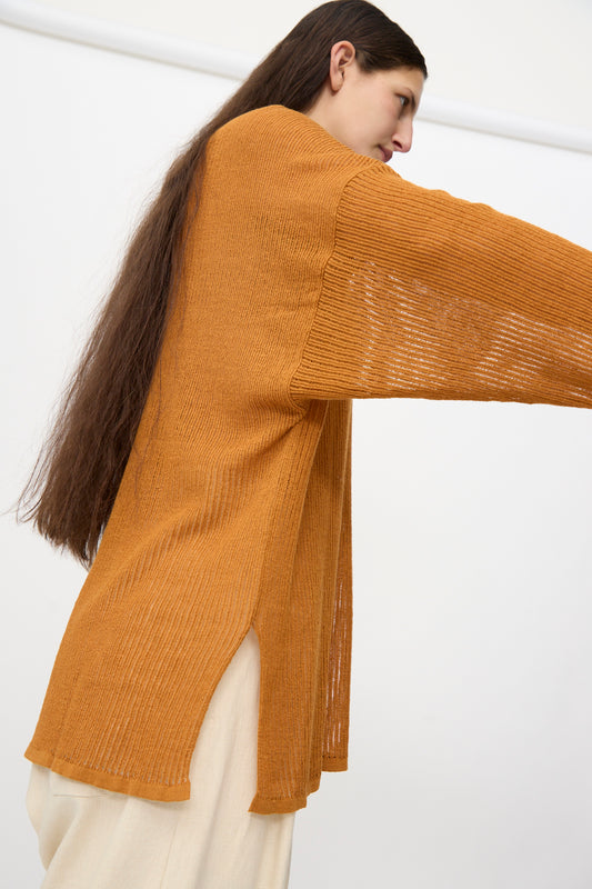 A person with long brown hair wears the Jan-Jan Van Essche Linen Cotton Mesh Knit #76 in Orange—an oversized, loose knit sweater with side slits—styled with cream pants and viewed from the side against a plain background.