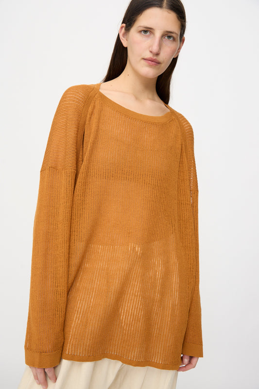 A woman wearing the Jan-Jan Van Essche Linen Cotton Mesh Knit #76 in orange, an oversized long-sleeve ribbed boatneck shirt, layered over a light skirt, stands against a plain white background.