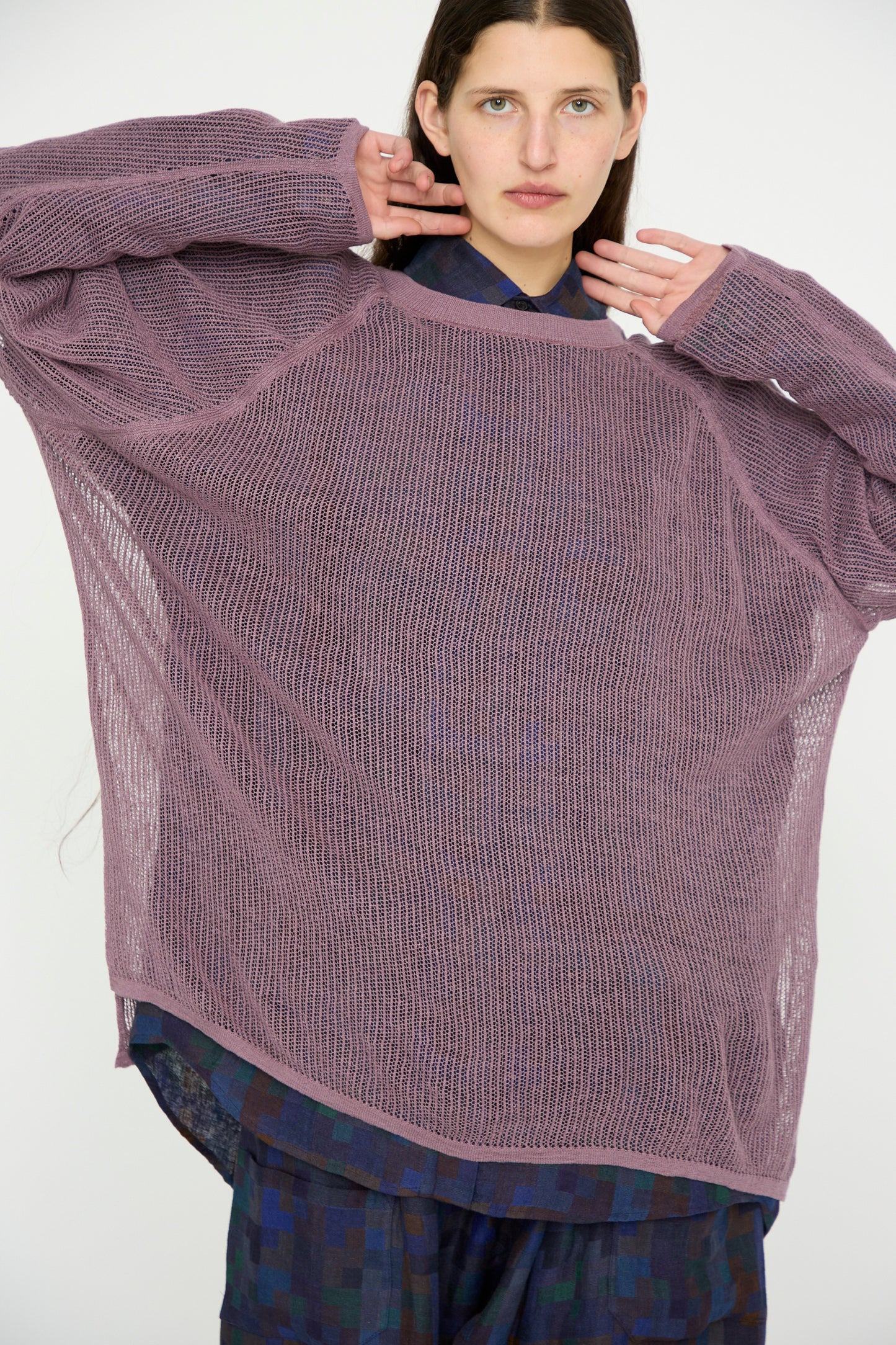 Against a plain backdrop, a person gracefully poses with hands near their face, wearing the Jan-Jan Van Essche Linen Cotton Yarn Knit #69 in Lavender, featuring loose sheer fabric and rib knit trims over a dark patterned outfit.