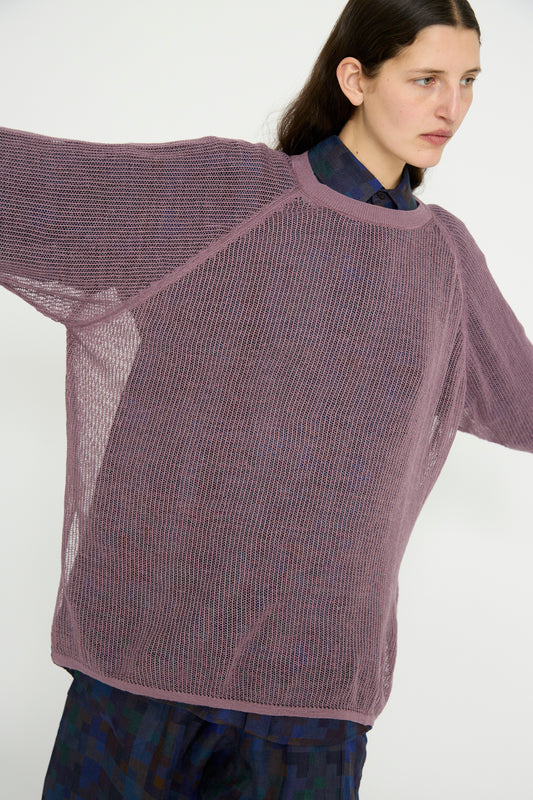 Wearing Jan-Jan Van Essche's Linen Cotton Yarn Knit #69 in Lavender, a model  extends their arms against a light backdrop. The oversized sweater features an intricate mesh ajour design and rib knit trims, layered over a dark plaid shirt.