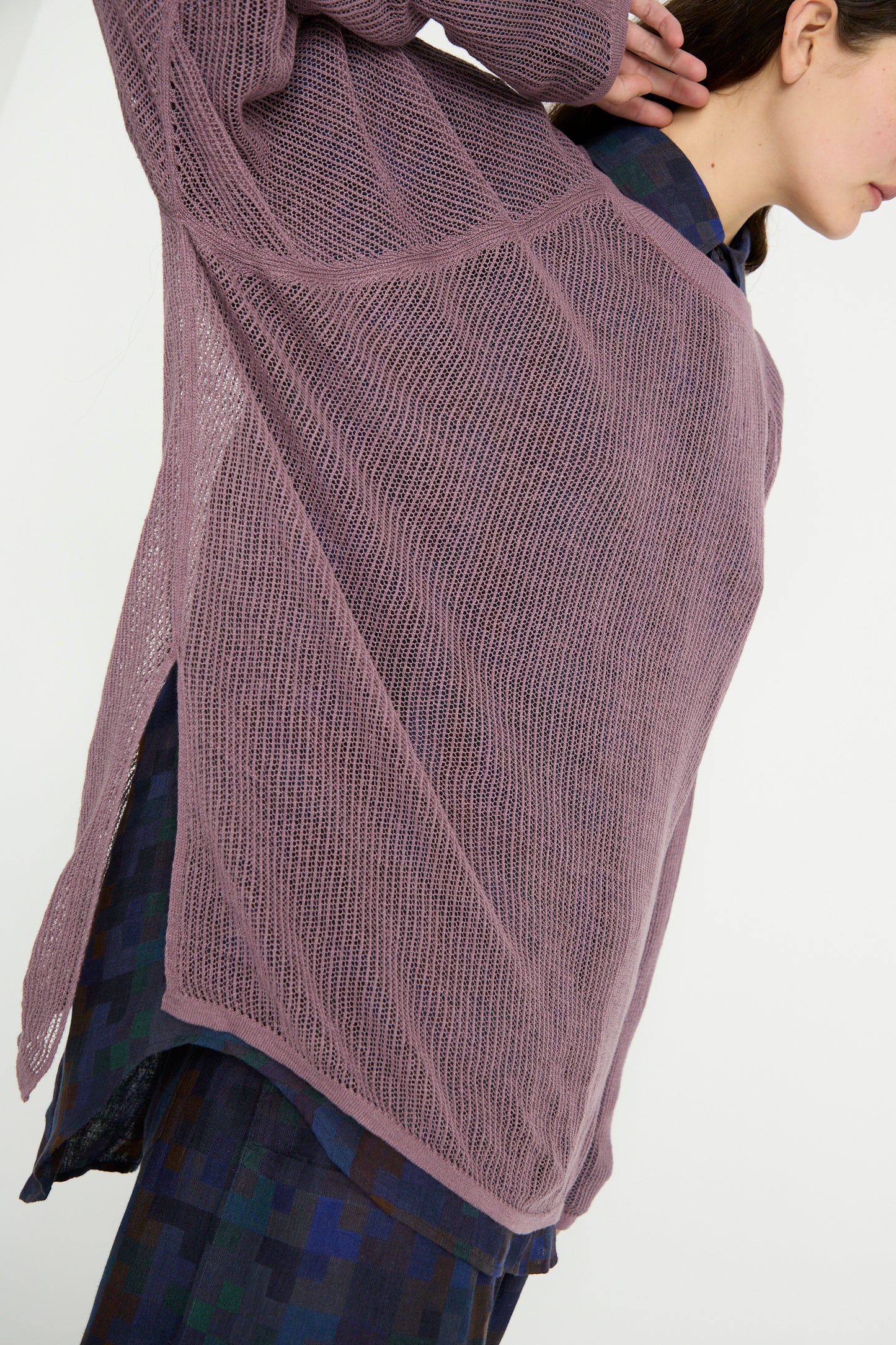 Draped in Jan-Jan Van Essche's Linen Cotton Yarn Knit #69 in Lavender, a person elegantly layers this oversized mauve sweater with intricate mesh ajour and chic rib trims over a blue-green patterned shirt, adding texture and sophistication to the ensemble.