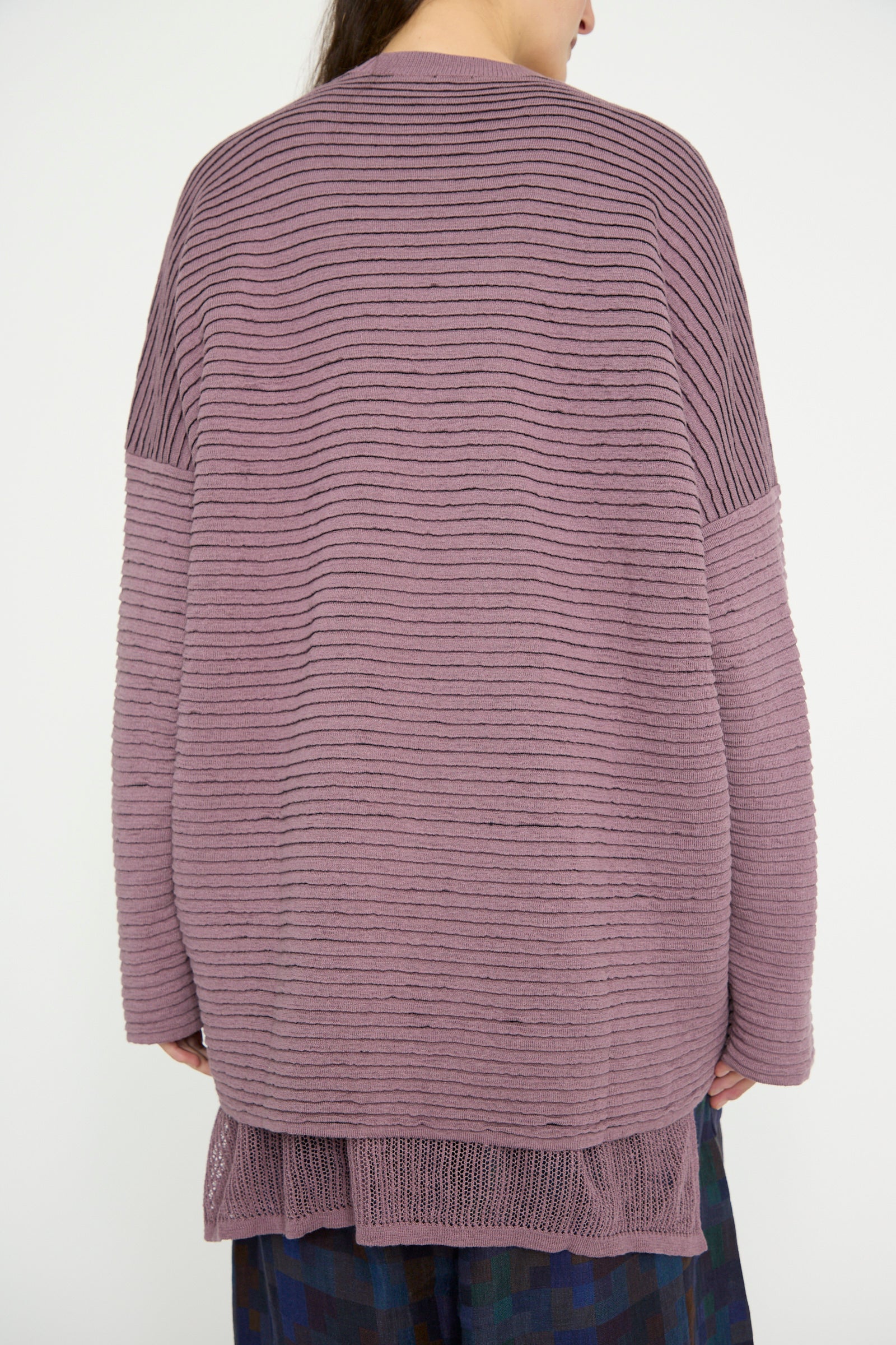 A person in an oversized Jan-Jan Van Essche Linen Cotton Yarn Knit #70 in Lavender turns away from the camera. The layered hem drapes over dark patterned pants against a plain backdrop.