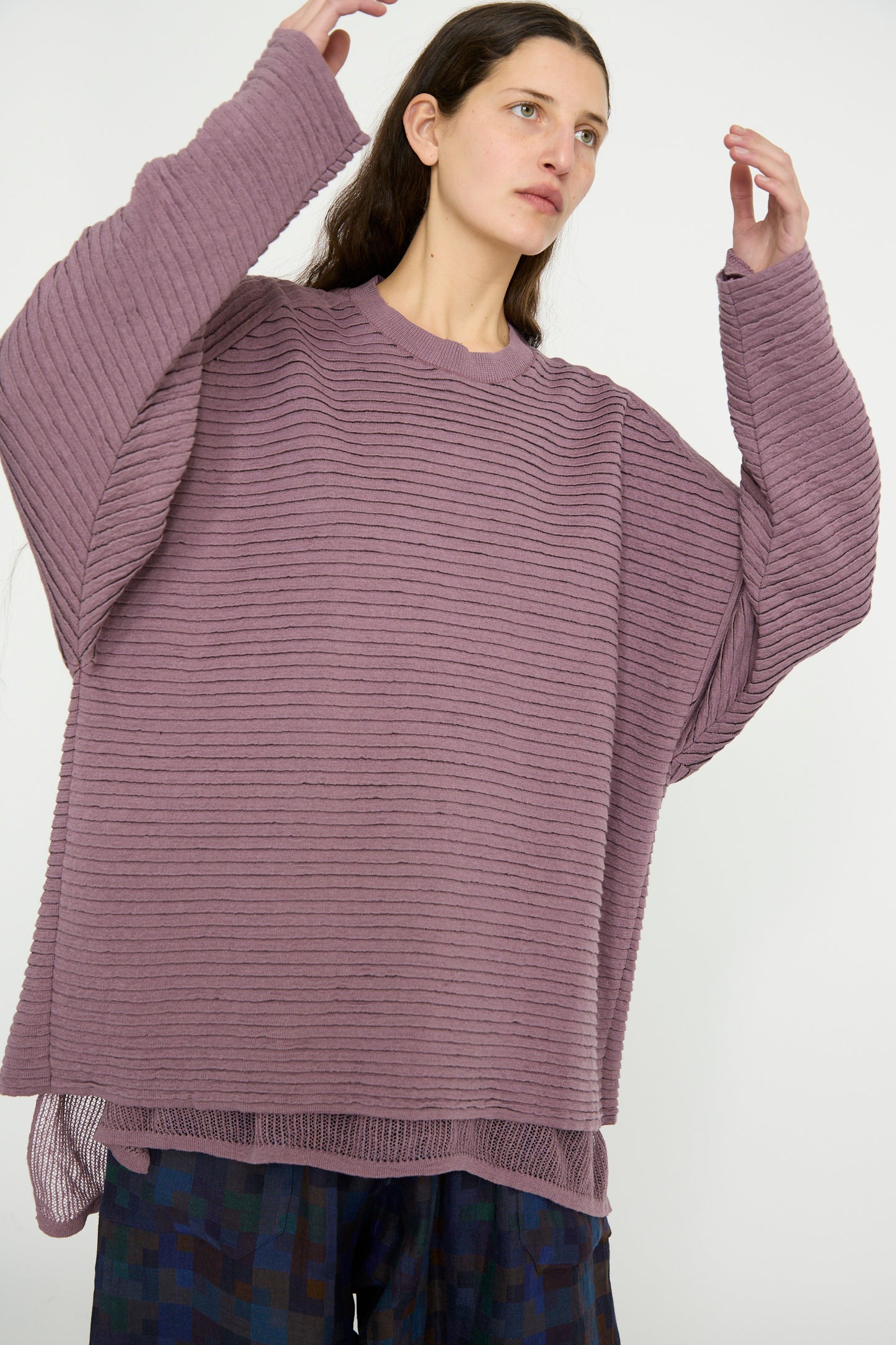 A person poses with arms raised against a plain background, wearing Jan-Jan Van Essche's oversized Linen Cotton Yarn Knit #70 sweater in Lavender paired with patterned pants.